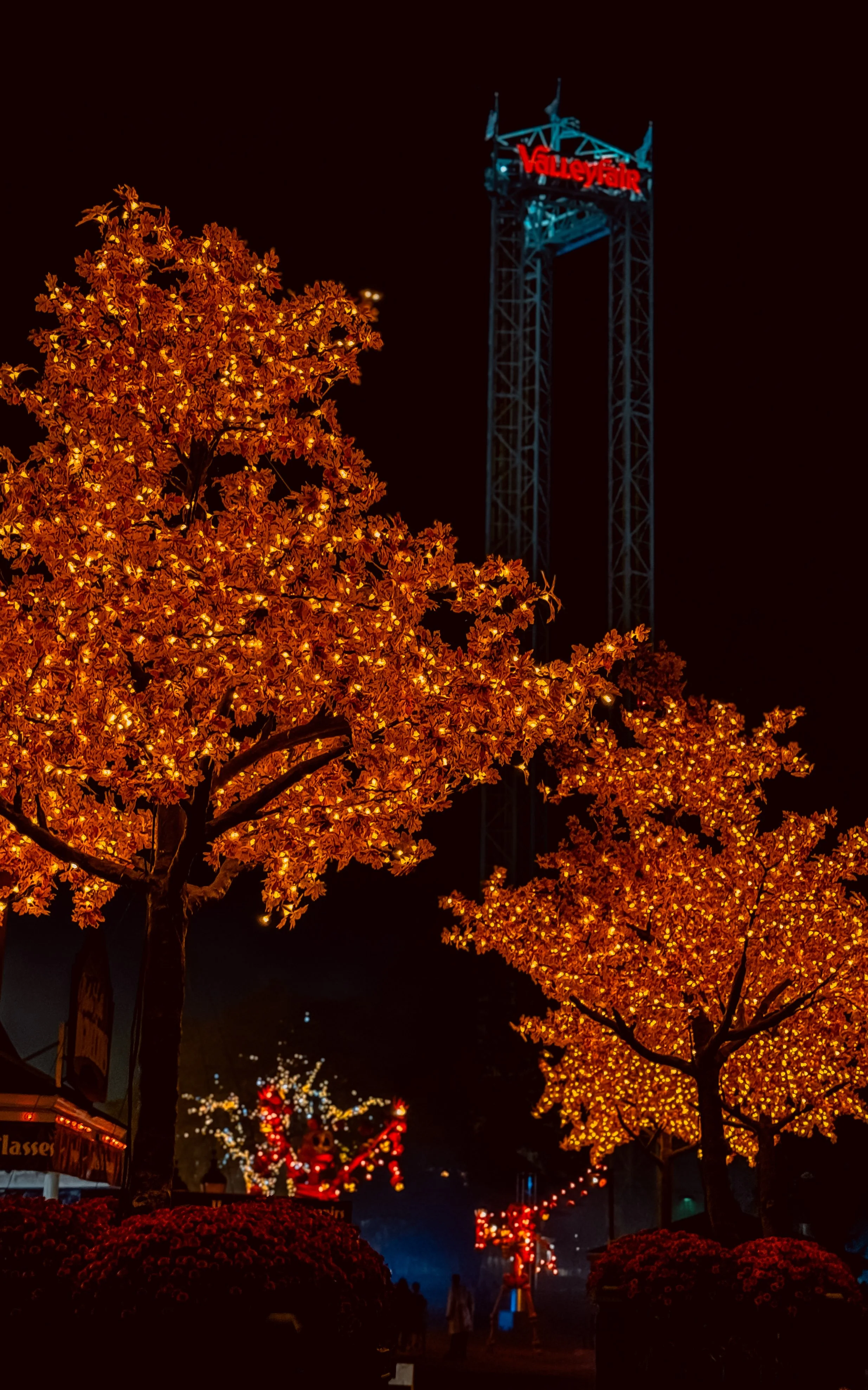 Night scene with illuminated orange and yellow trees, a ValuCheck sign on a tall tower, and a festive display in the background.
