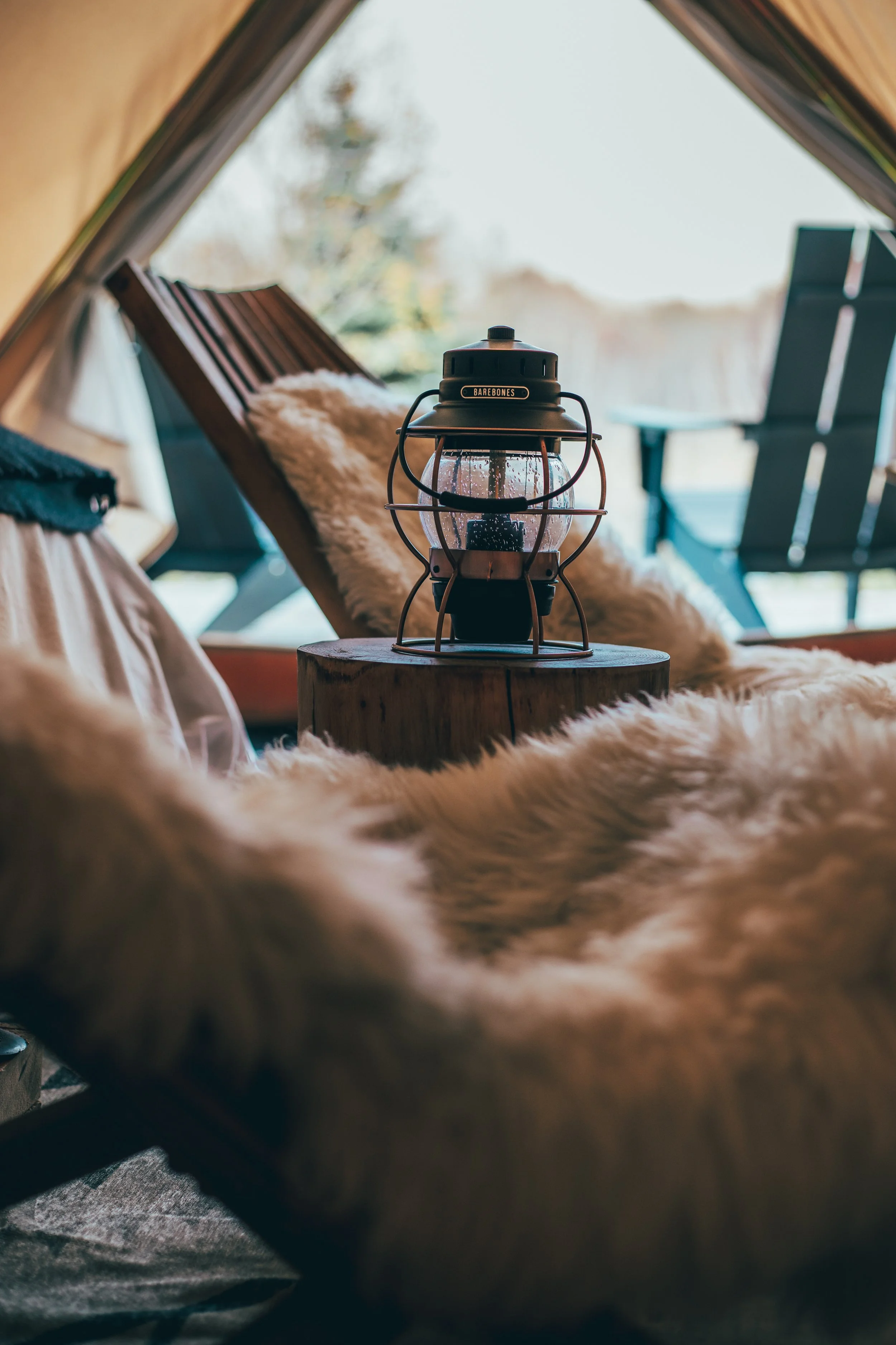 Inside a cozy camping tent with a Sheepskin rug, a lantern on a wooden stump, and outdoor chairs visible through the tent opening.