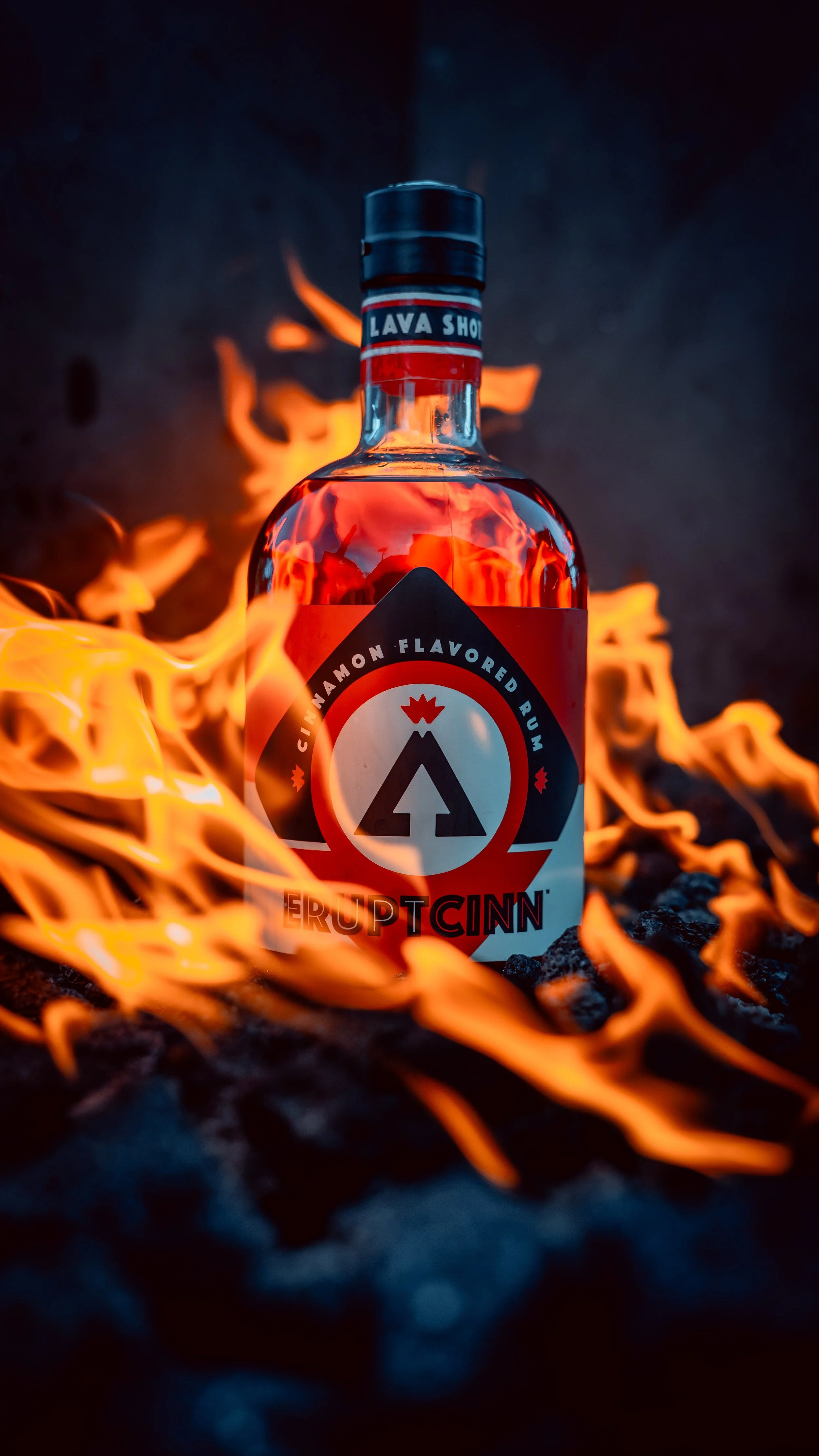 A bottle of flavored rum labeled 'ERUPT Cinn' is standing on black rocks, surrounded by flames, with a dark background.