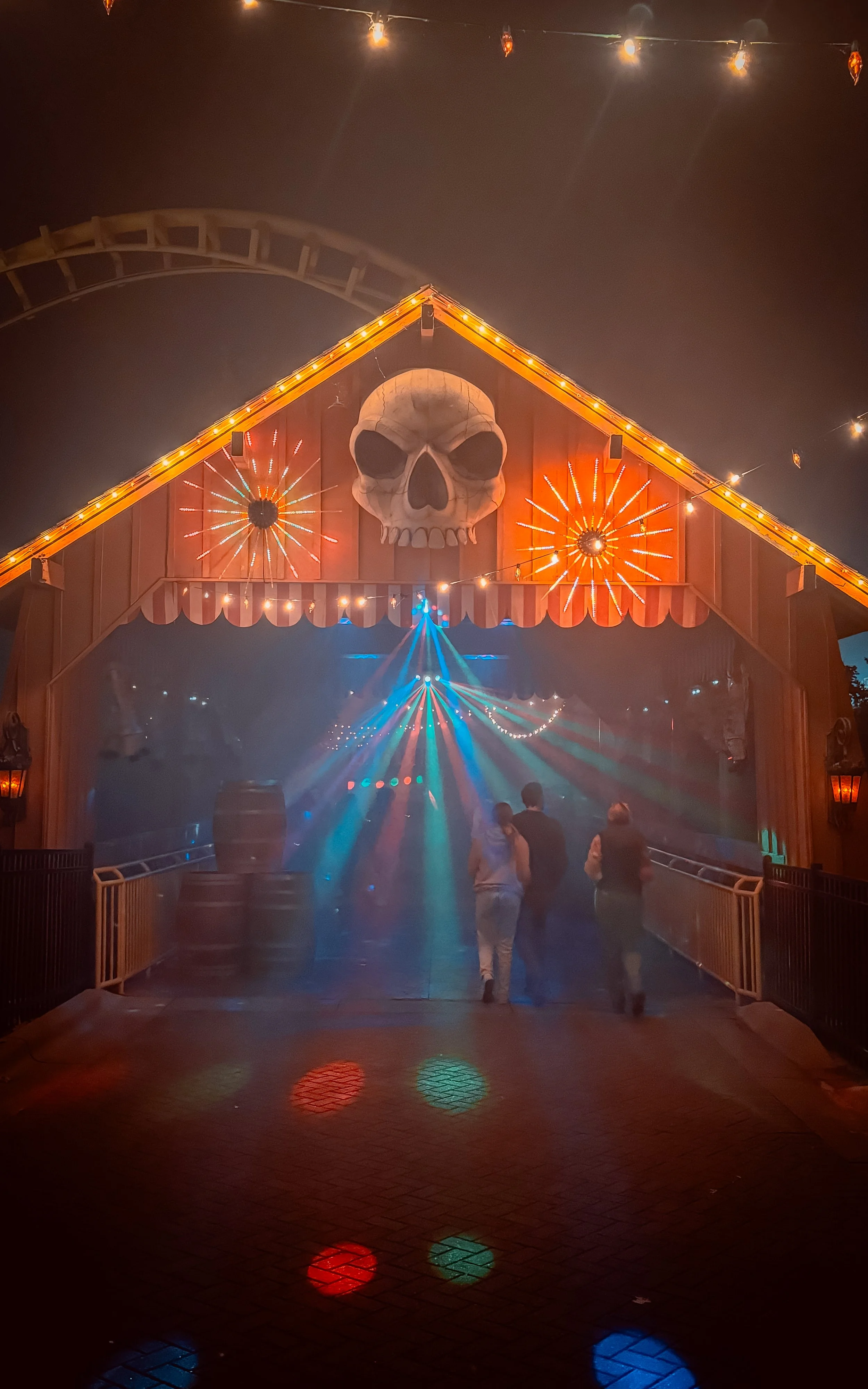 Nighttime scene at a haunted house attraction with a large skull decoration on the building's peaked roof, illuminated by orange lights and firework-like decorations. There are laser lights and fog effects inside, with three people walking toward the