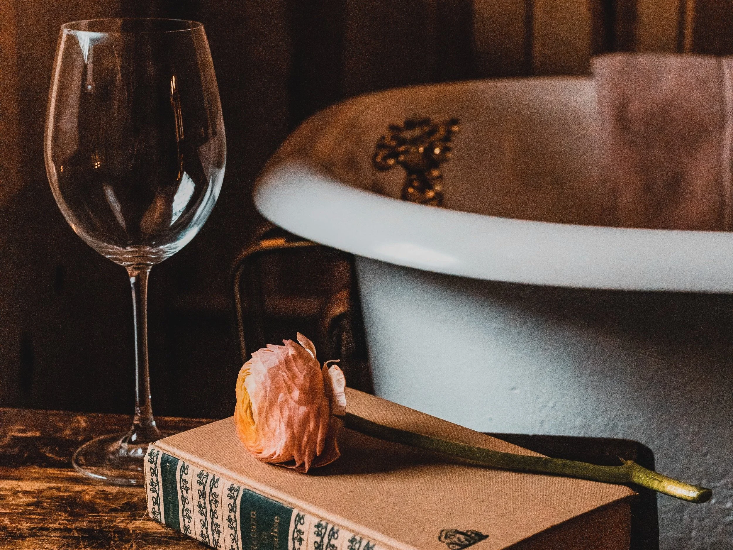 A wine glass, a pink flower, and a closed book with decorative edges on a wooden table, with a bathtub in the background.