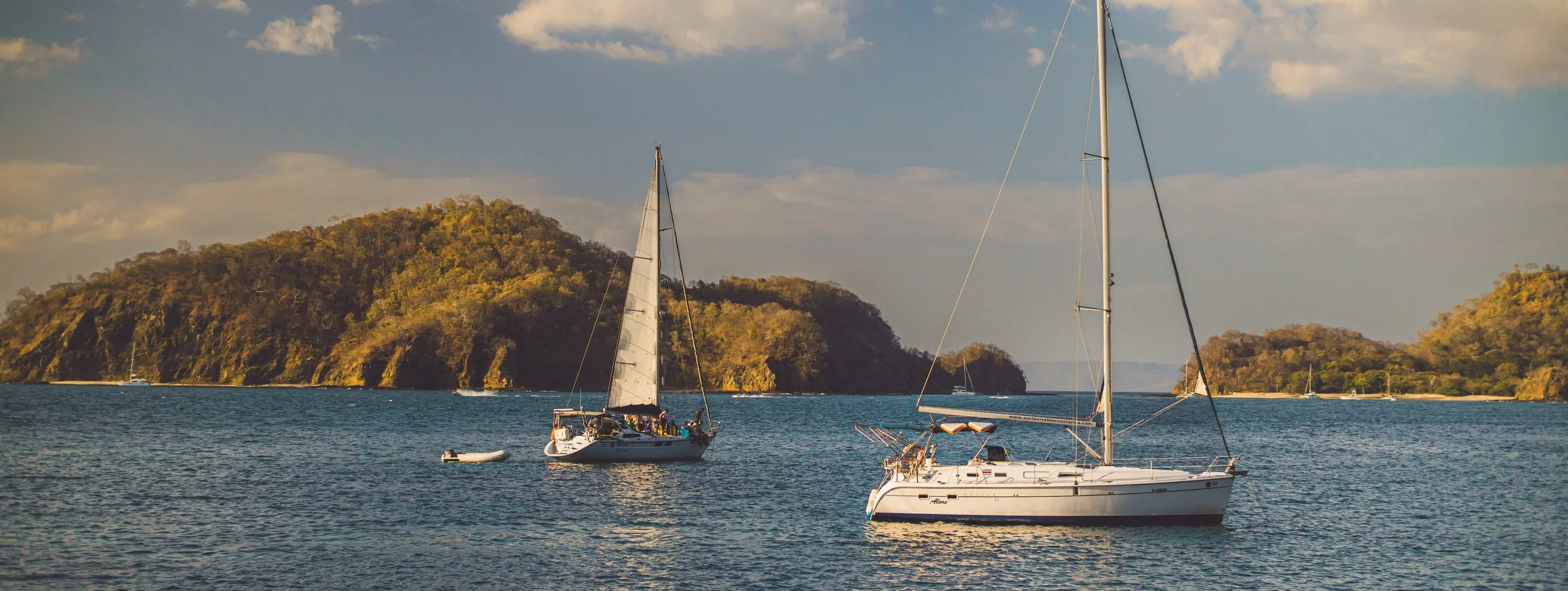 Sailboats anchored in a calm bay with forested islands in the background under a partly cloudy sky.