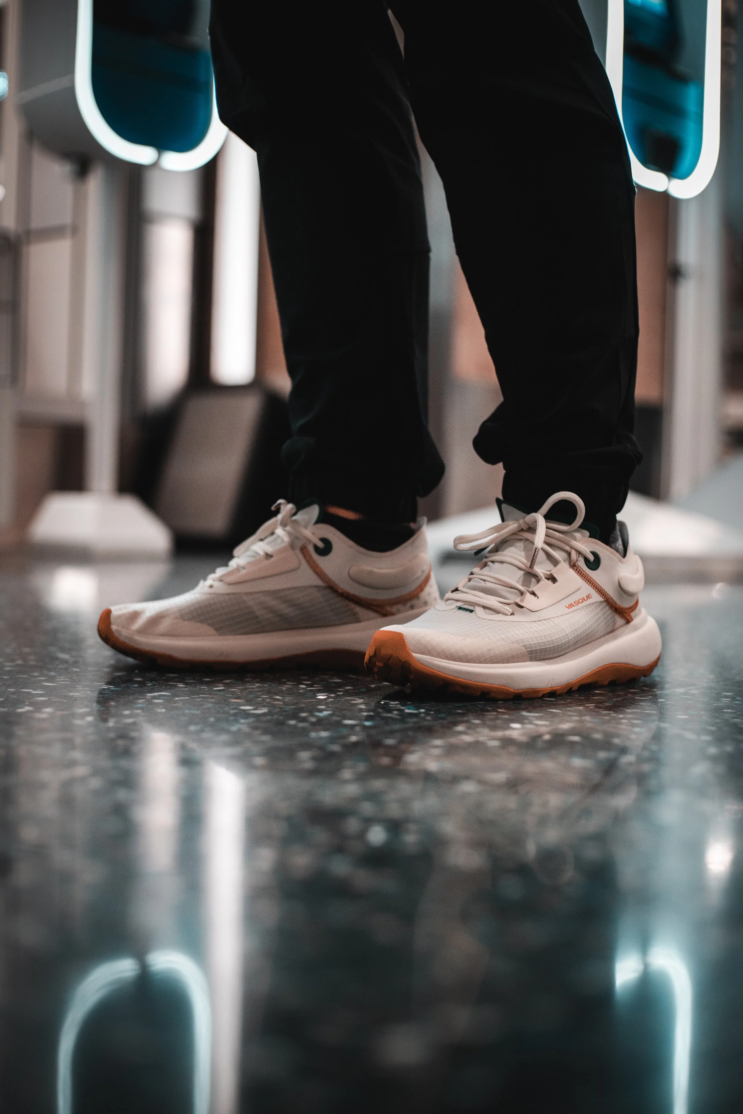 Close-up of a person's feet wearing white and orange running shoes, standing on a dark, reflective floor in what appears to be an airport or train station, with seating and structural elements in the background.