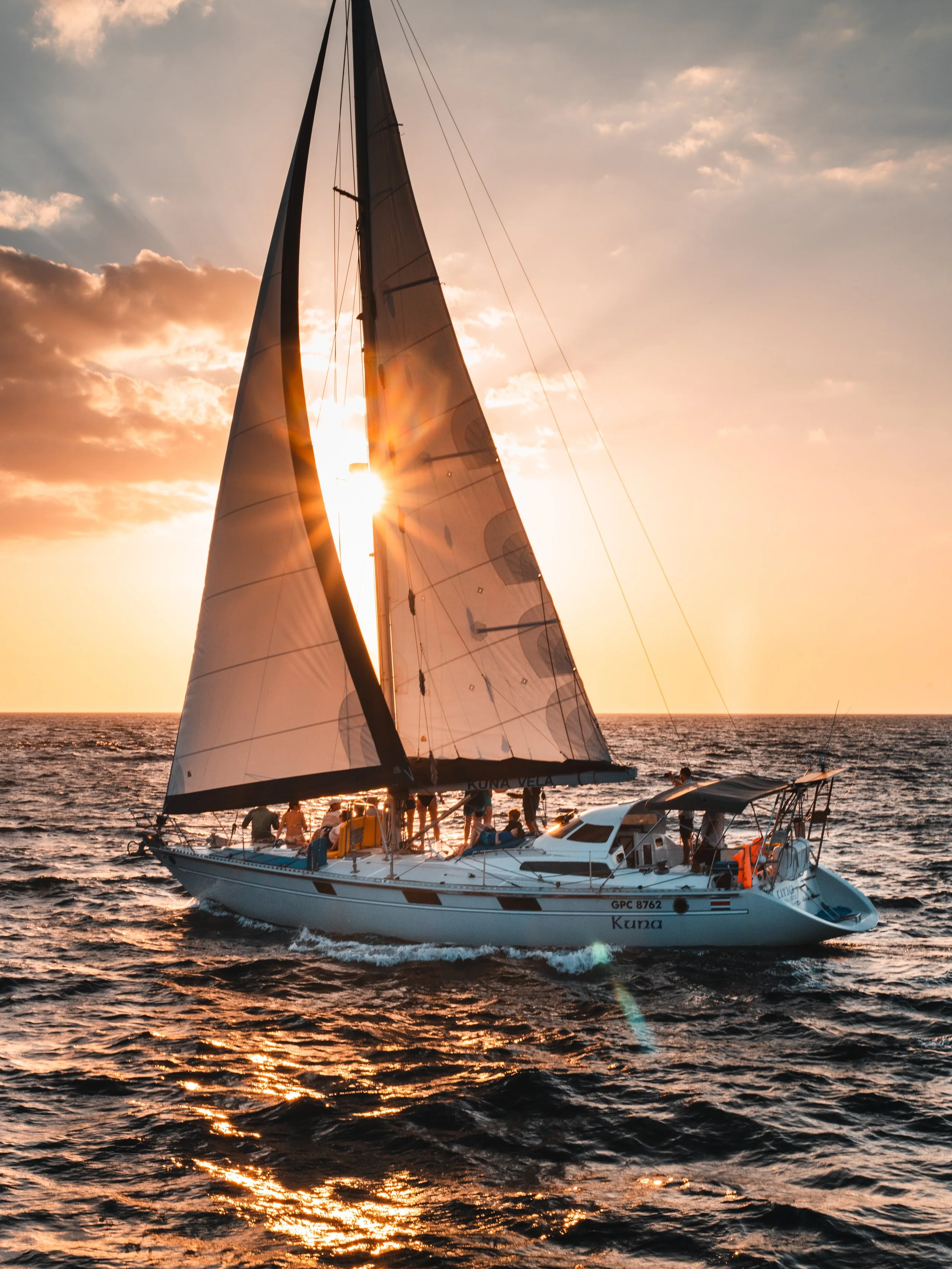 A sailboat with large black and white sails on the ocean during sunset, with the sun partially behind the sails and people on deck