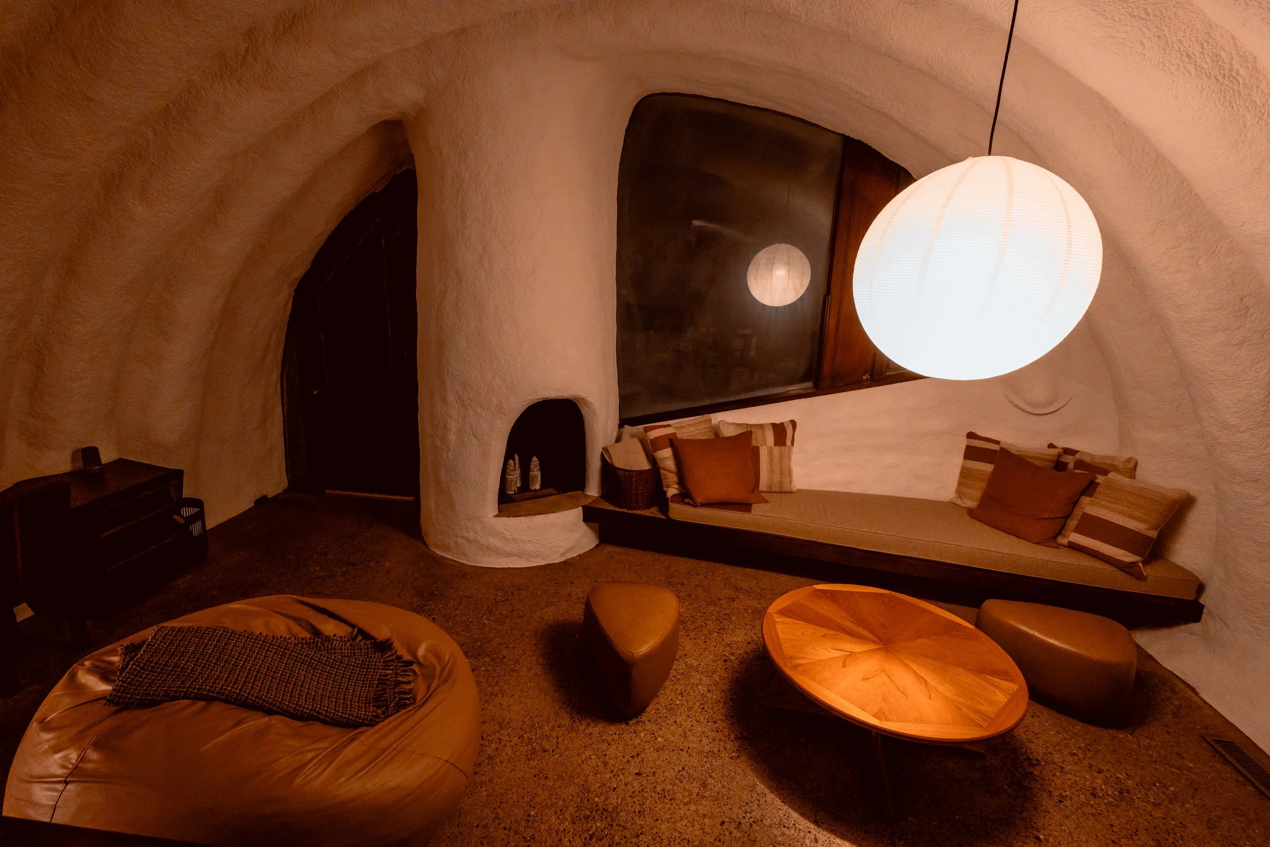 Cozy living room with white textured curved walls, large round paper lantern hanging from ceiling, cushioned window seat with pillows, wooden coffee table, leather pouf, and small heater, illuminated by warm ambient lighting.