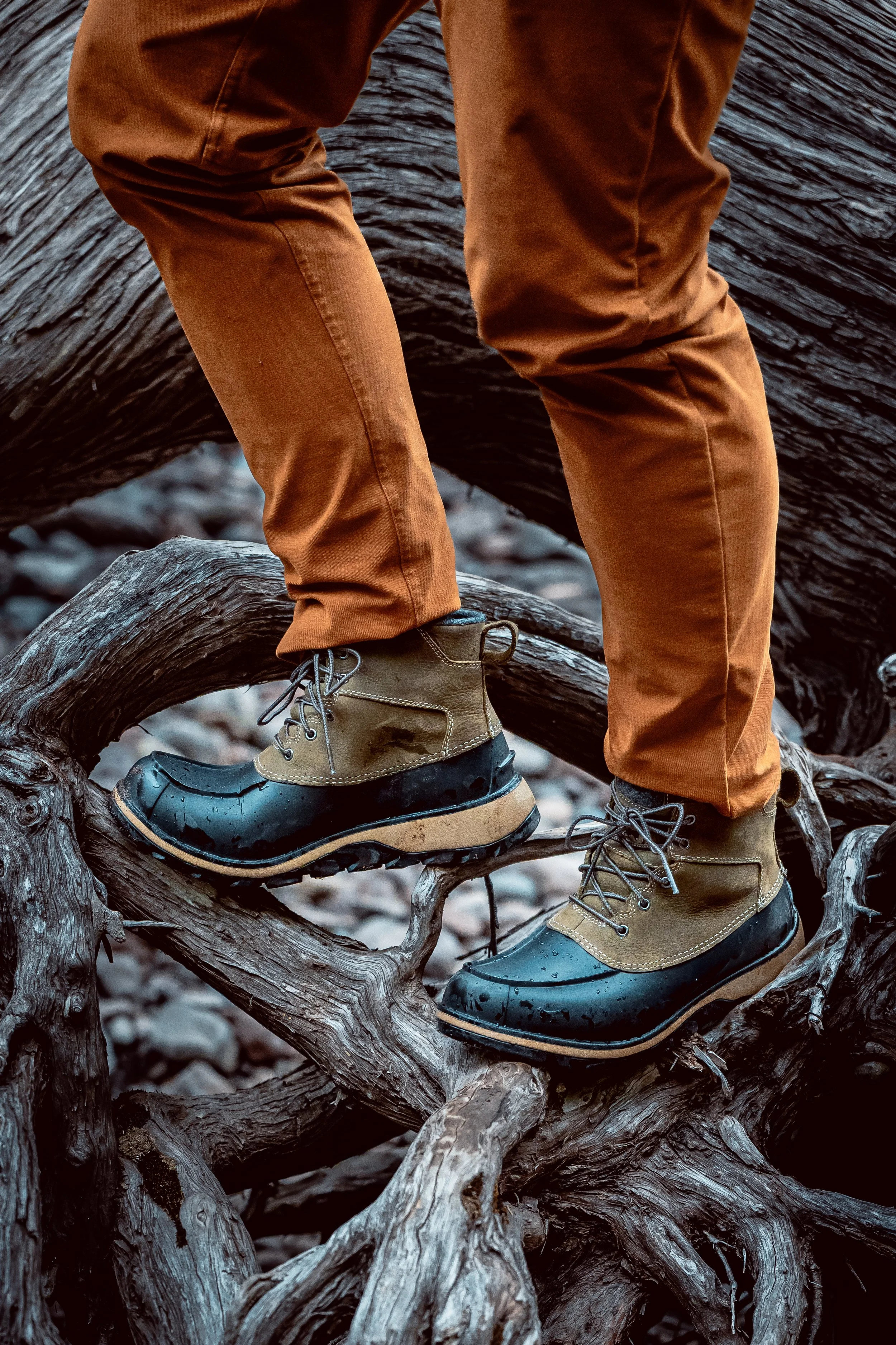 A person wearing waterproof boots and brown pants balancing on a tree branch near water.