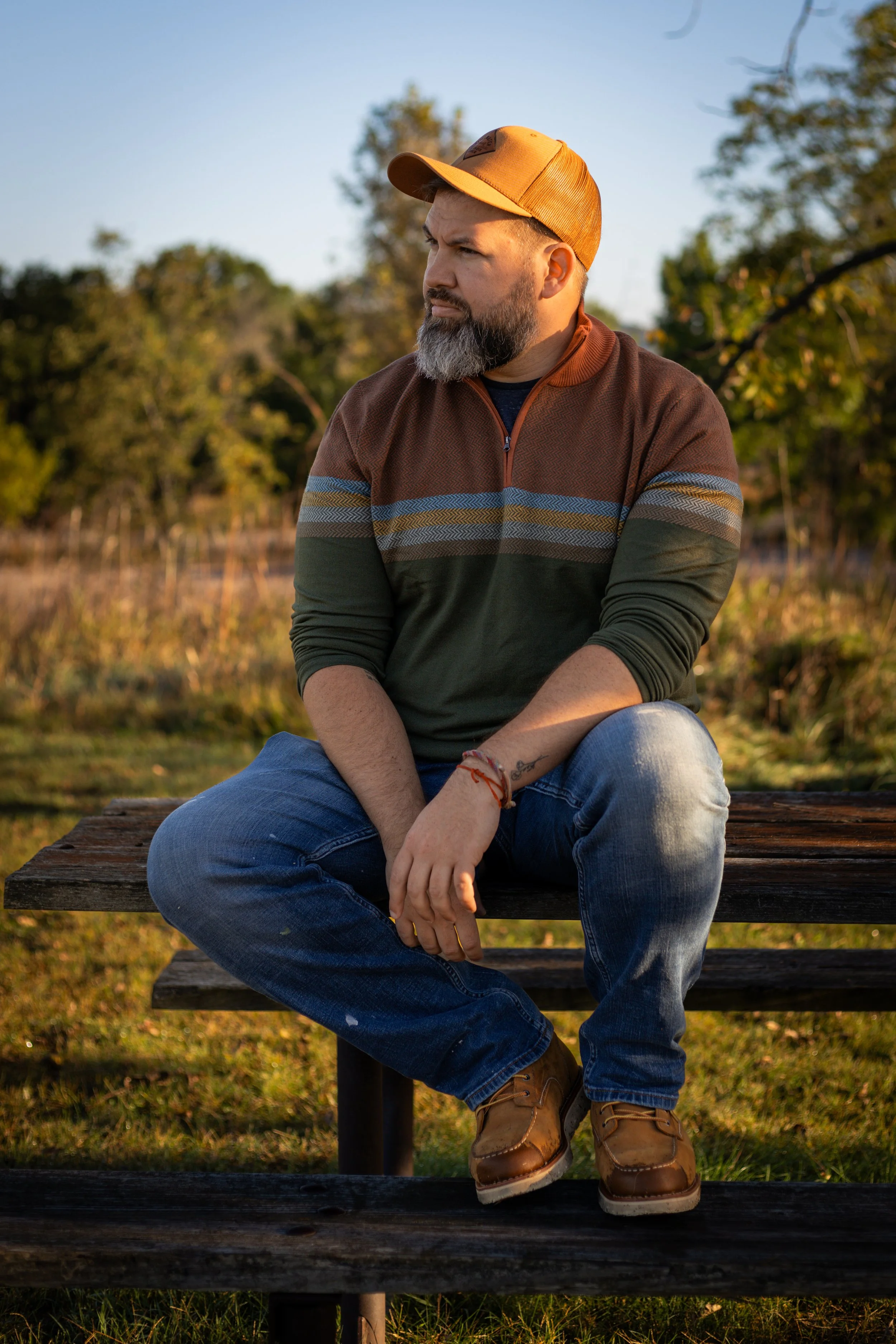 A bearded man wearing a tan cap, a colorful striped sweater, jeans, and tan boots sitting on a wooden bench outdoors during sunset, with trees in the background.
