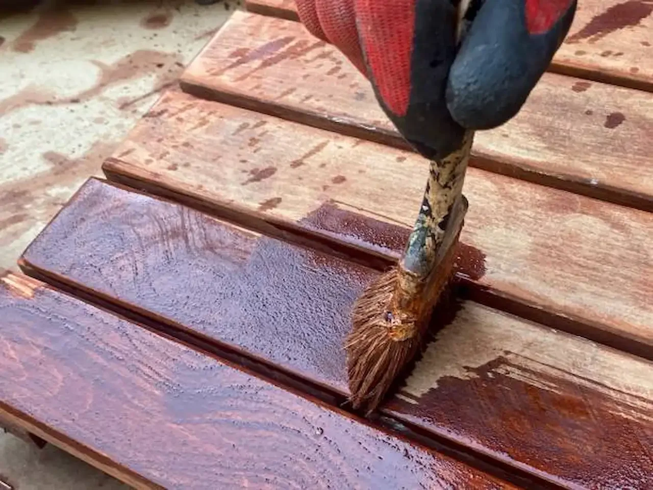 Close-up of a gloved hand applying dark wood stain with a paintbrush onto wooden planks, showing freshly coated glossy surface and unfinished boards beside it.