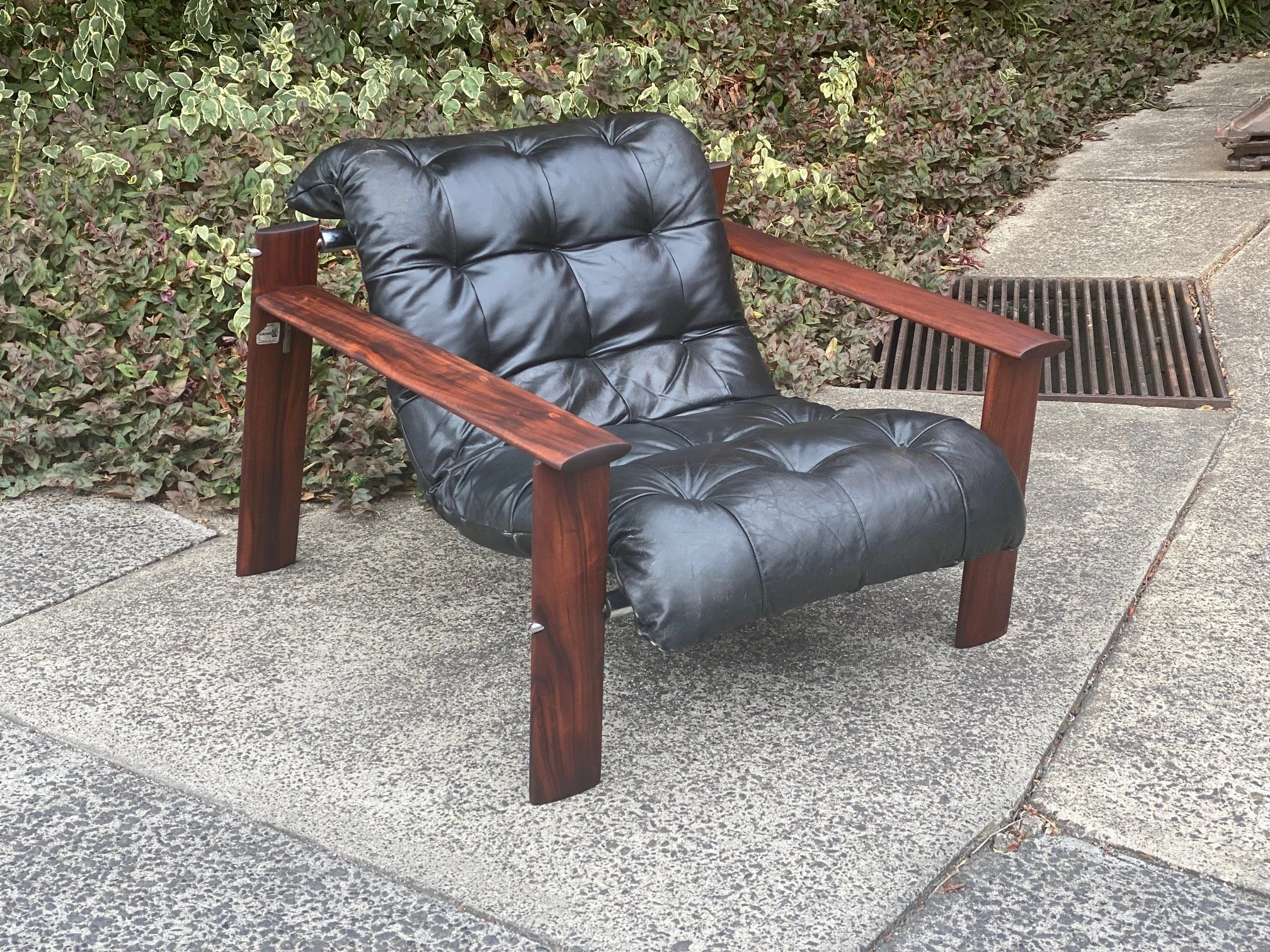 Outdoor black leather armchair with wooden armrests and legs on a concrete sidewalk next to a hedge and a drainage grate.