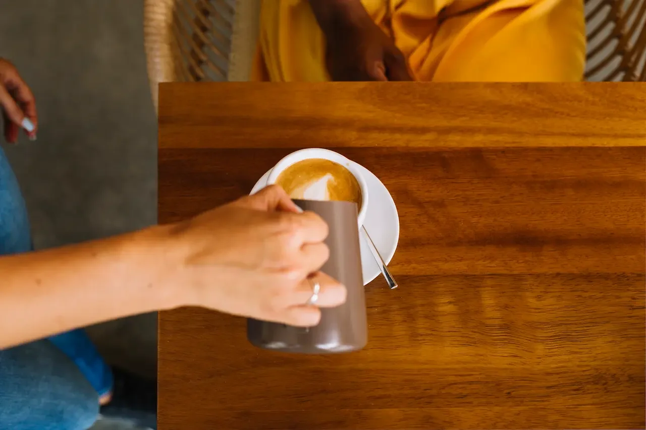 Close-up of a woman’s hand holding a coffee mug over a wooden table with a cappuccino on a saucer.