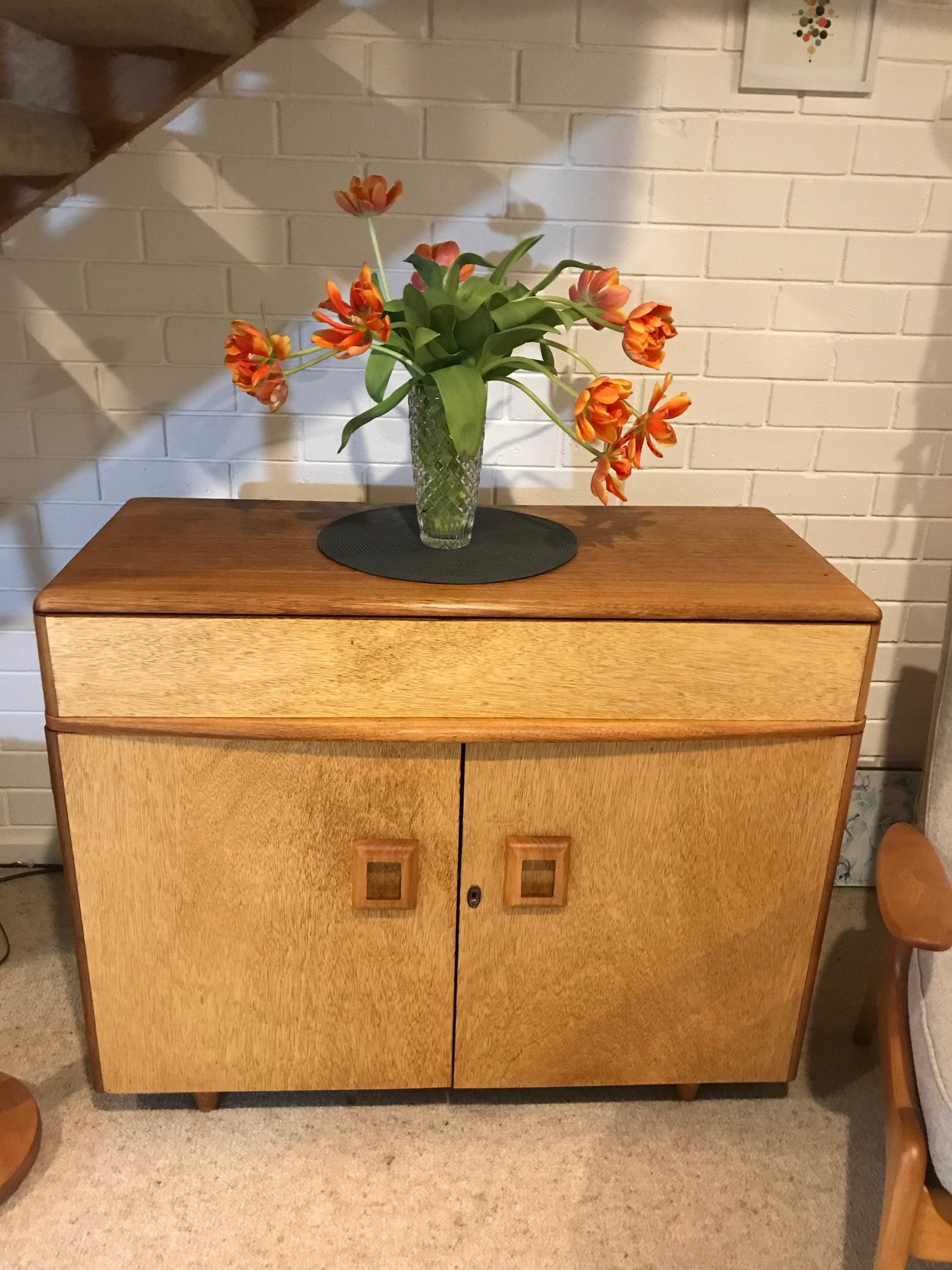 Wooden cabinet with a textured glass vase of orange tulips on top, placed on a black circular placemat, against a white brick wall.