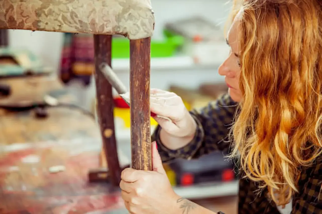 Close-up of a woman carefully restoring a vintage wooden chair leg, applying treatment with a small tool in a workshop setting.