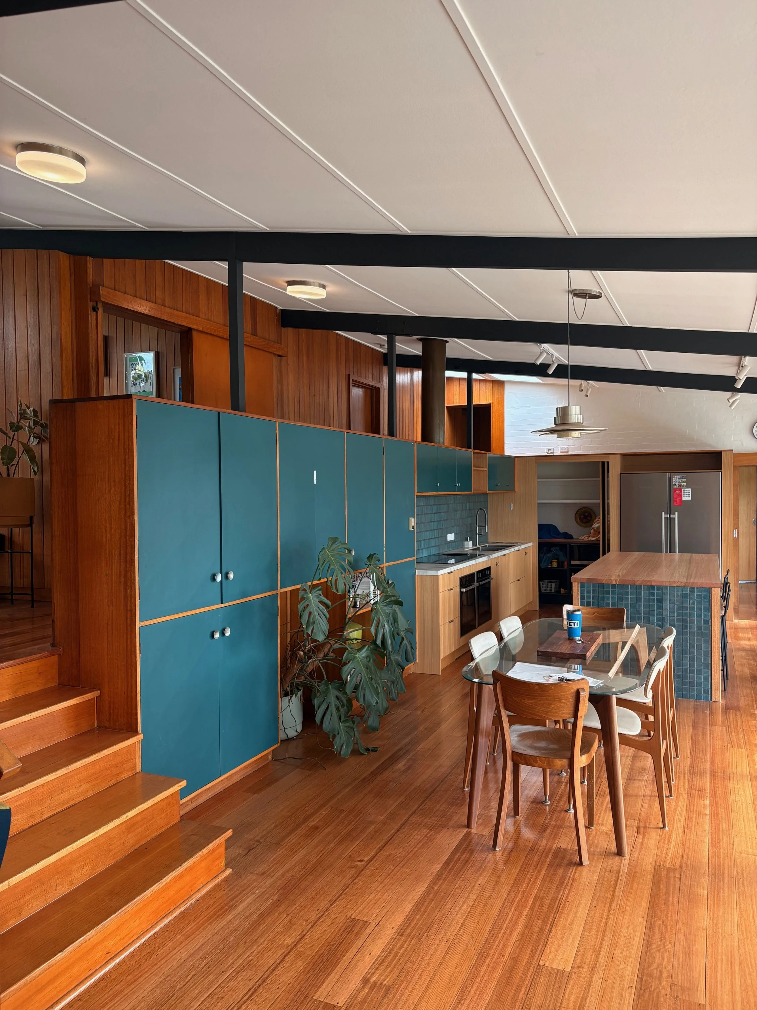 Indoor kitchen and dining area with wooden floors, teal cabinets, wooden dining table with four white and wooden chairs, and a kitchen counter with a sink and oven. There are large green plants and modern ceiling lights.