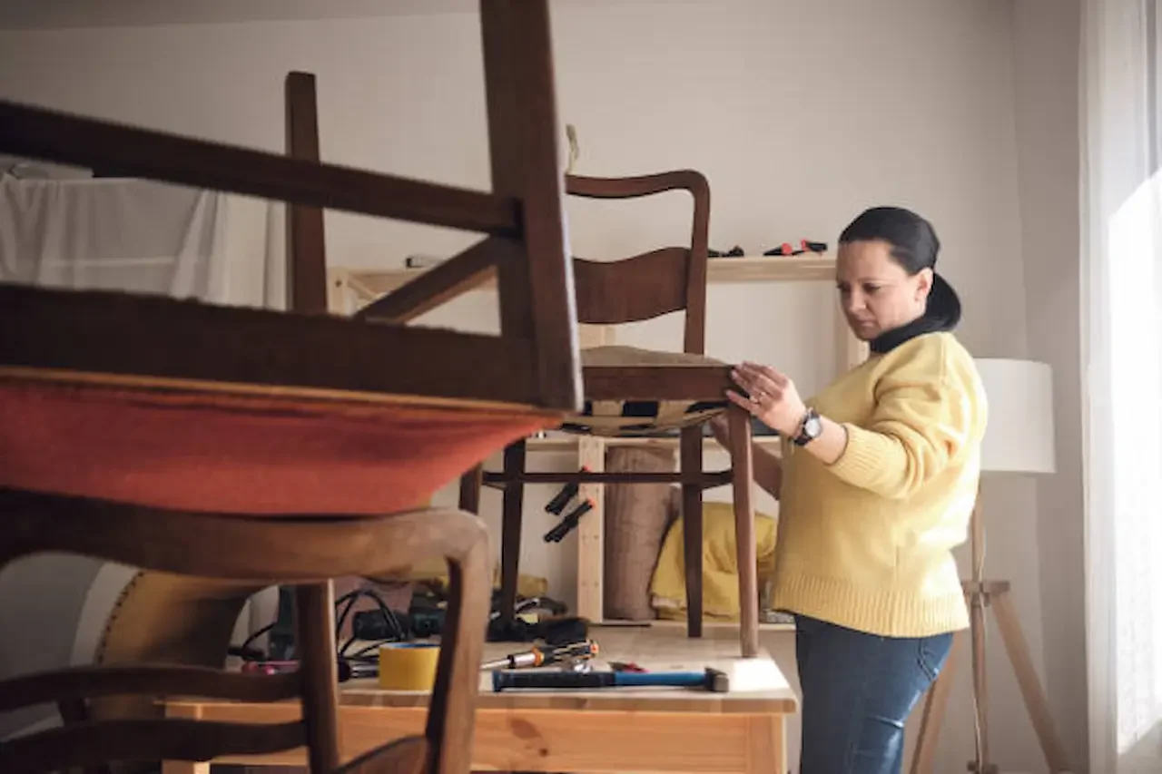 Woman carefully repairing a wooden chair in a home workshop, with tools visible on the table and another chair positioned upside down in the foreground.