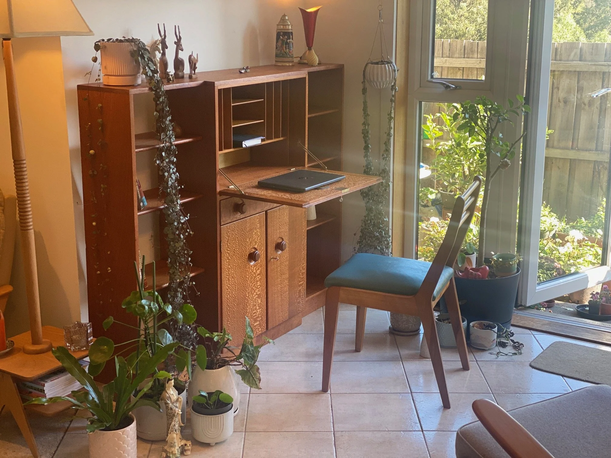 Sunlit living room corner with a wooden desk, a wooden chair with a teal seat, potted plants, and decorative items on a cabinet, next to open glass door leading outside to a garden.
