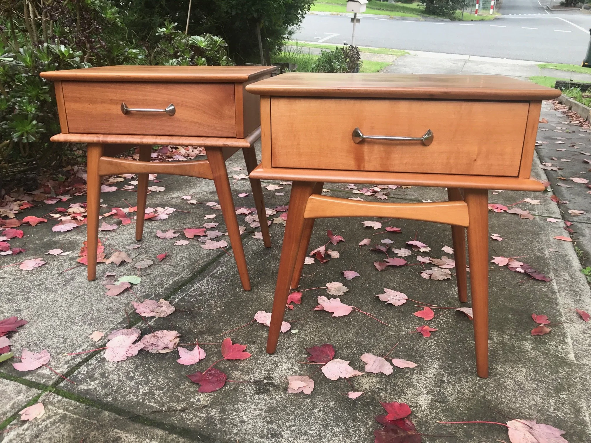 Pair of wooden nightstands with drawers, placed outdoors on a concrete sidewalk with fallen red and pink leaves, greenery, and a street in the background.