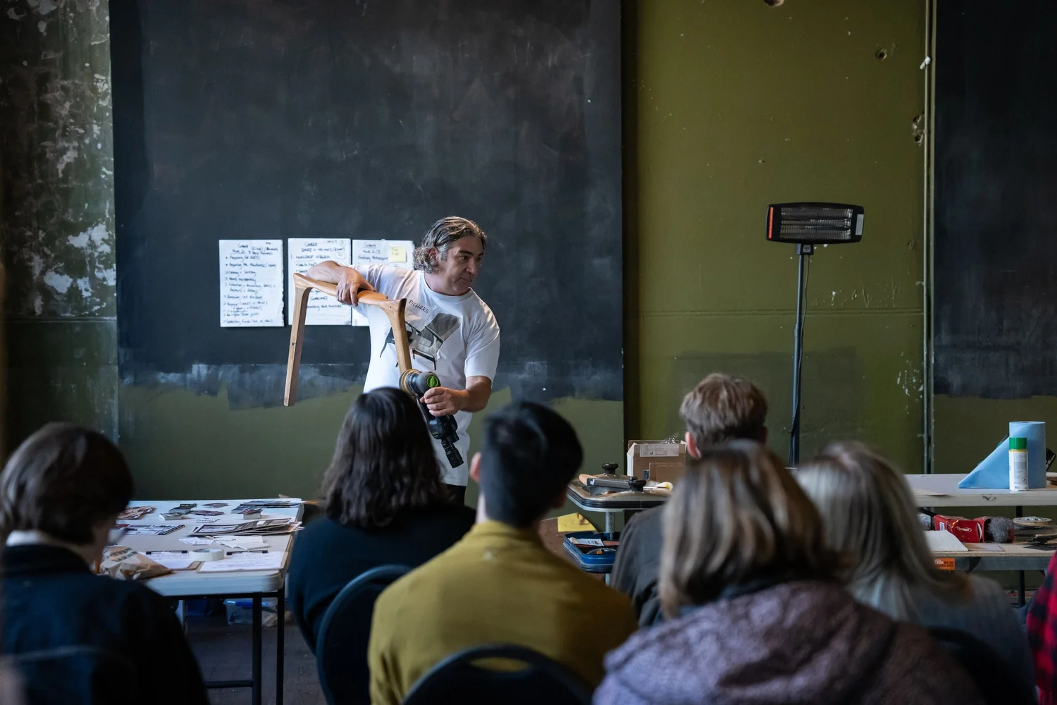 A man with shoulder-length hair holds a chair and microphone while speaking to a group of students in a classroom or workshop setting. The students are seated at tables with various materials, and a blackboard with papers and notes is behind the man. A heater and miscellaneous objects are visible in the background.