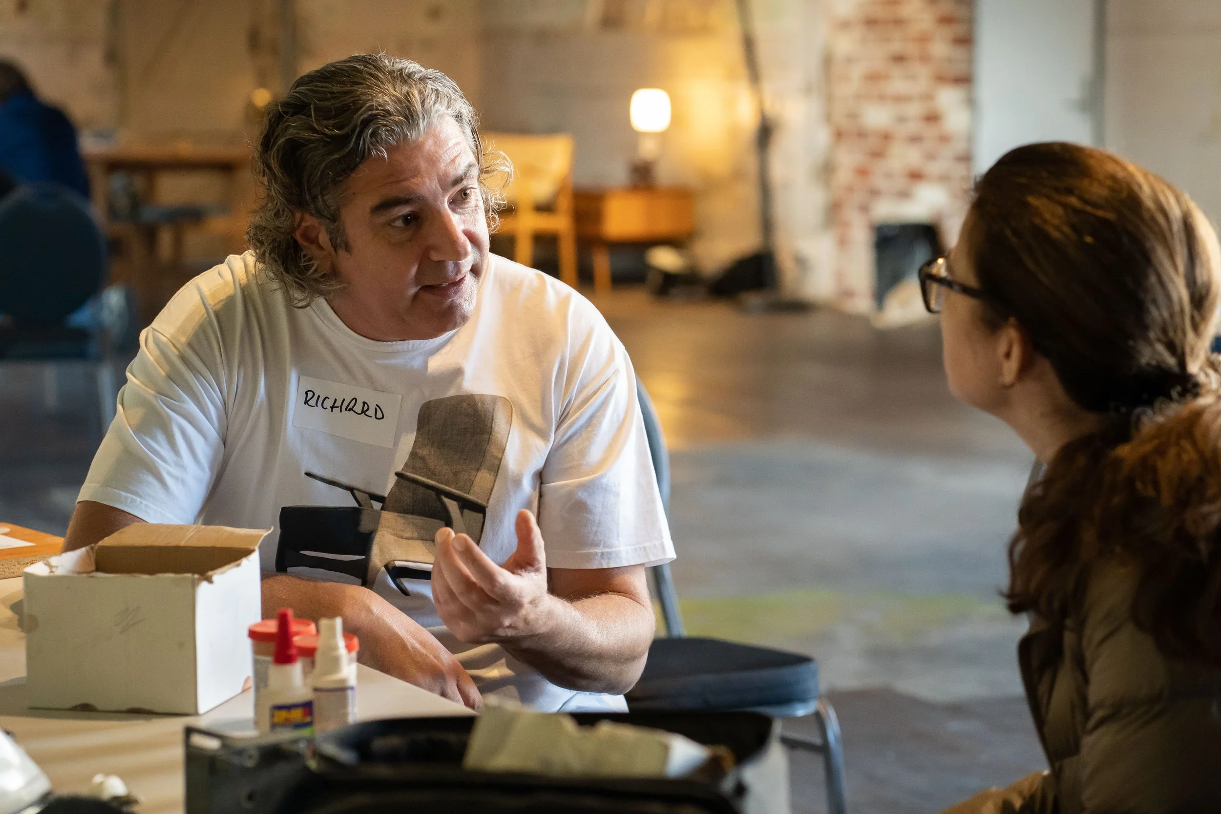 A man with curly gray hair and a name tag that says 'Richard' is talking to a woman with glasses and long brown hair in a casual indoor setting with warm lighting.
