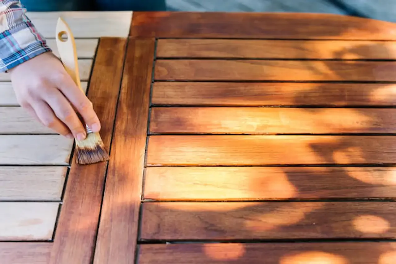 Close-up of a person applying wood stain with a small brush to an outdoor wooden table, showing the contrast between freshly stained dark wood and lighter unfinished slats in sunlight.