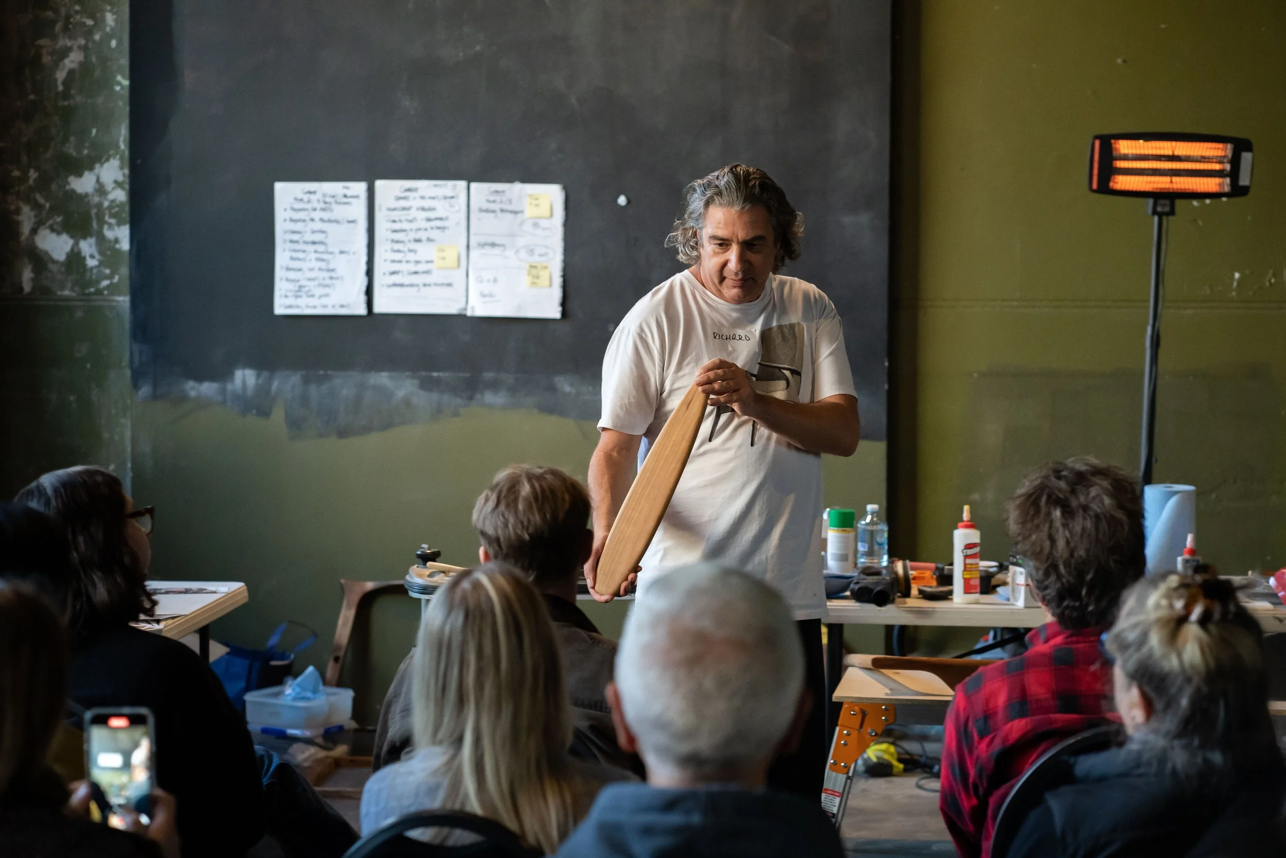 Man demonstrating a wooden skateboard to a seated audience in a workshop or classroom setting.