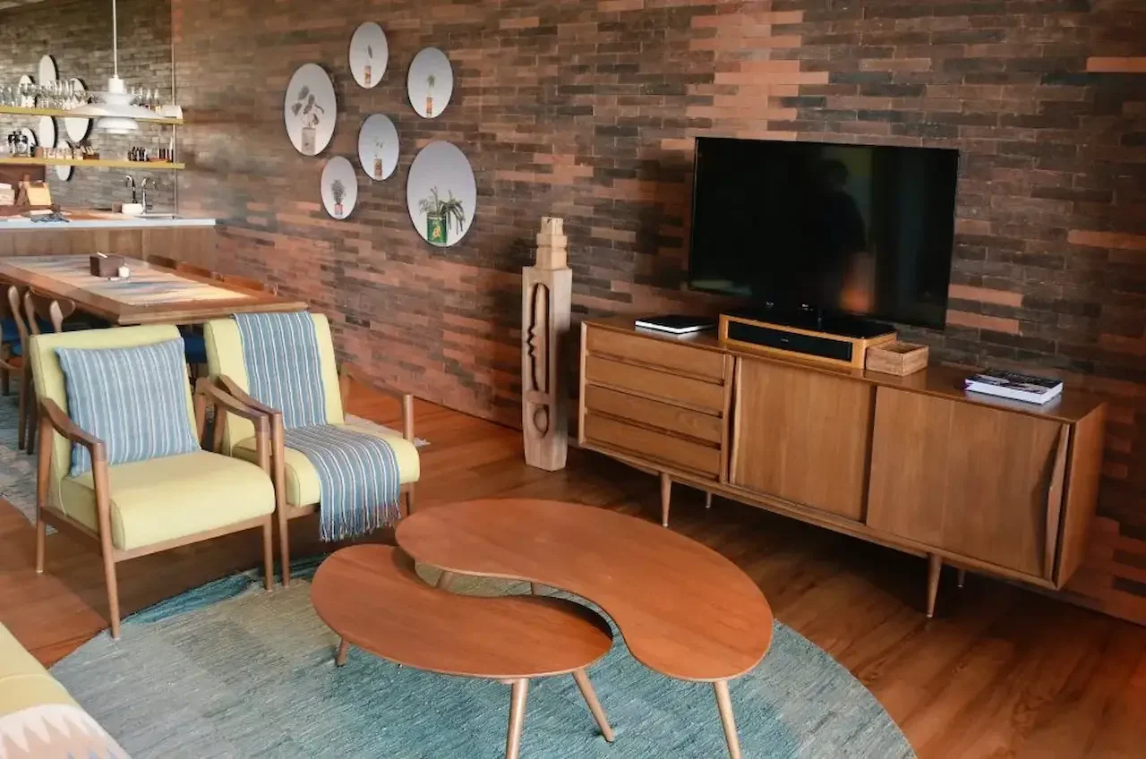 Mid-century modern living room featuring a teak sideboard with television, sculpted wooden coffee table, upholstered armchairs, and a brick accent wall with decorative mirrors.