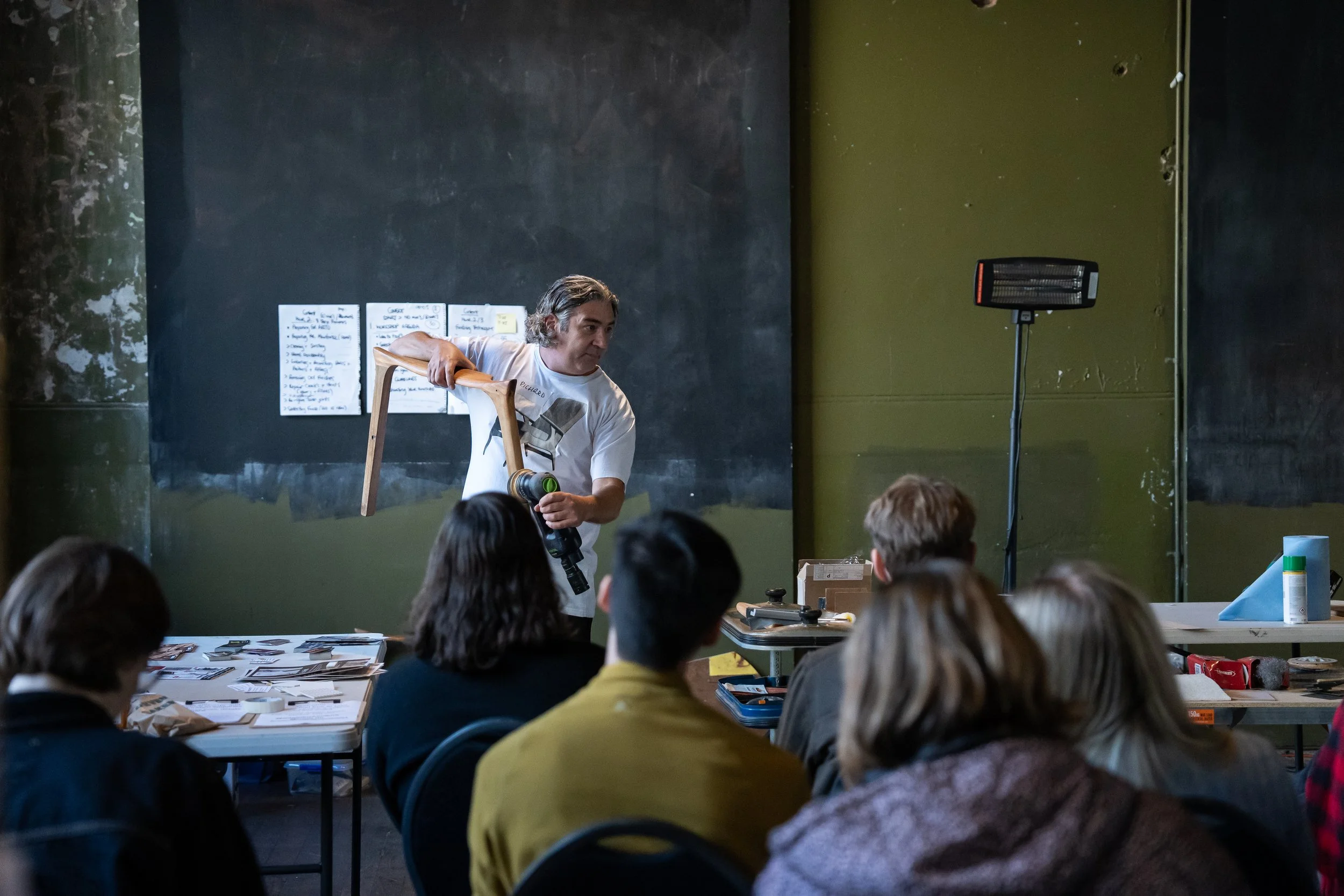 A man demonstrating using a small sledgehammer on a wooden chair in front of a seated audience in a workshop or class setting.