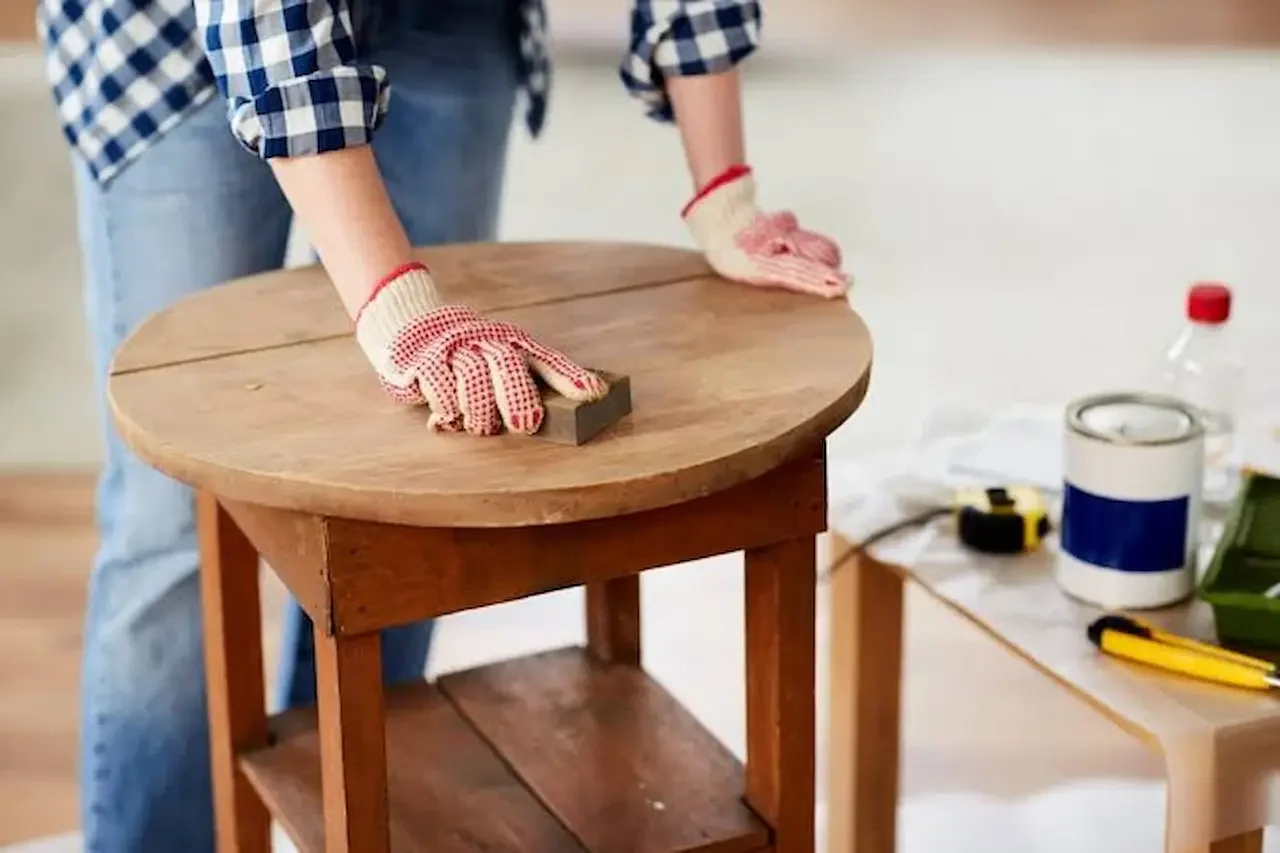 Person sanding a round wooden table while wearing red and white gloves, with tools and supplies on a nearby table.