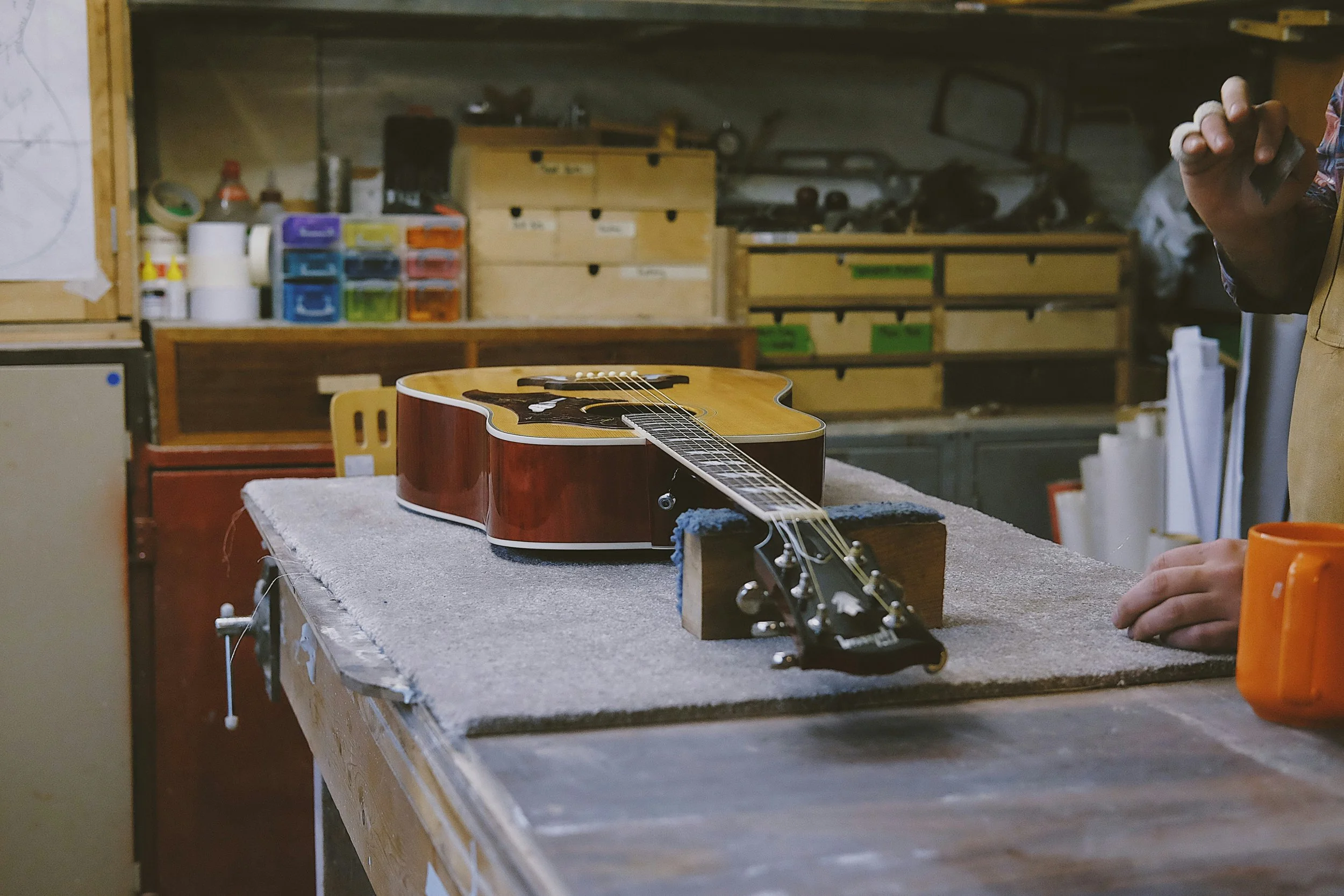 An acoustic guitar is on a worktable in a workshop, with a hand holding a tool on the right side of the image and various storage containers and tools in the background.