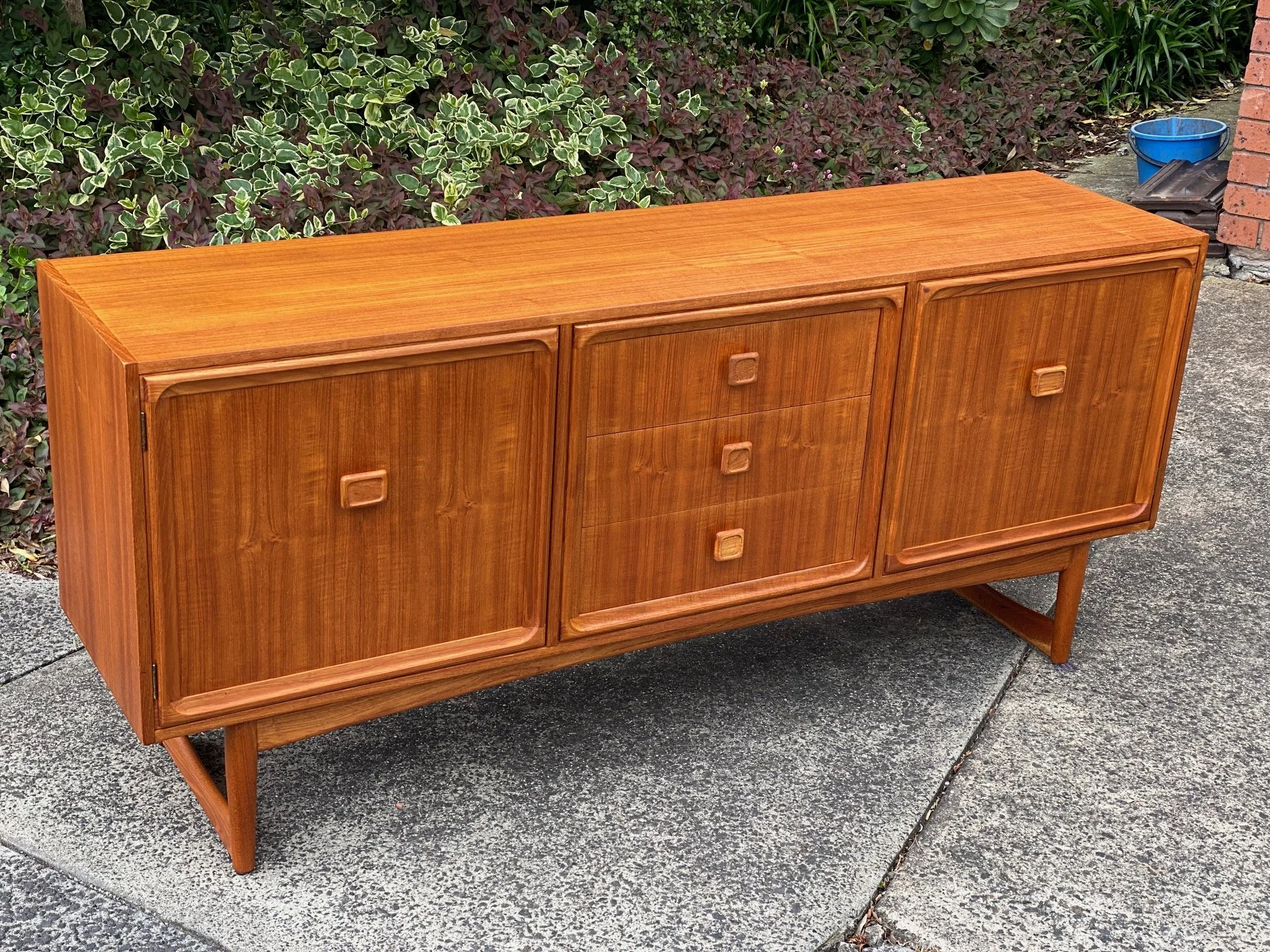 A wooden sideboard with sliding doors and three central drawers, set on a concrete outdoor surface near some greenery and a brick wall.