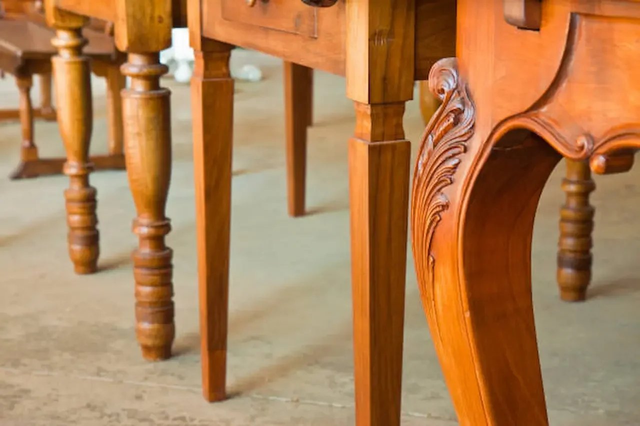 Close-up of polished wooden table legs featuring turned spindle designs and decorative carved detailing on one leg, set on a light-colored floor.