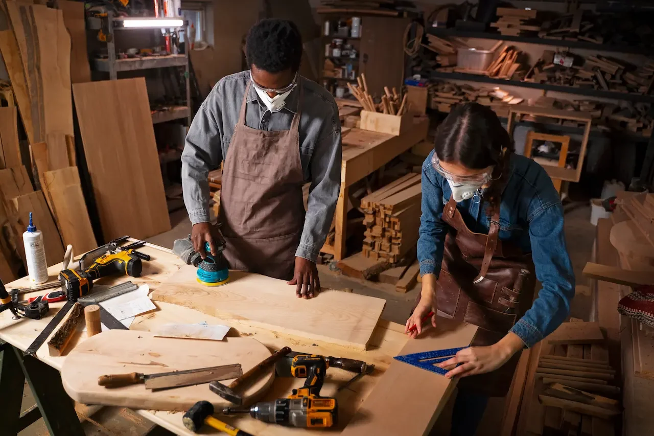 Two people working in a woodworking shop, sanding a large wooden piece. Both are wearing protective masks and aprons, with tools and supplies on the worktable.