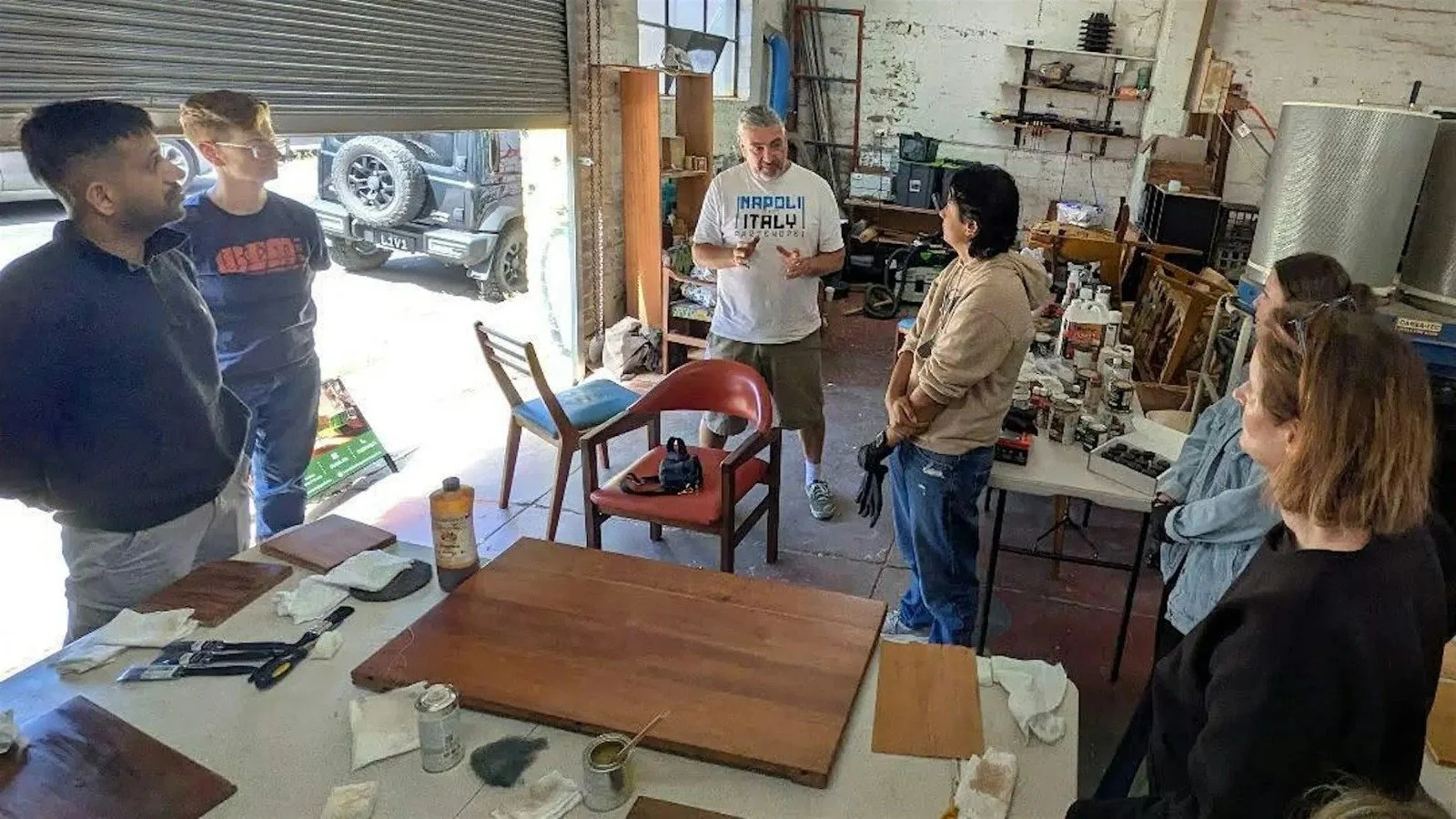 Group of people in a workshop or garage, one person speaking to others. The workspace has tables, tools, paints, and storage shelves, with a garage door open to the outside.