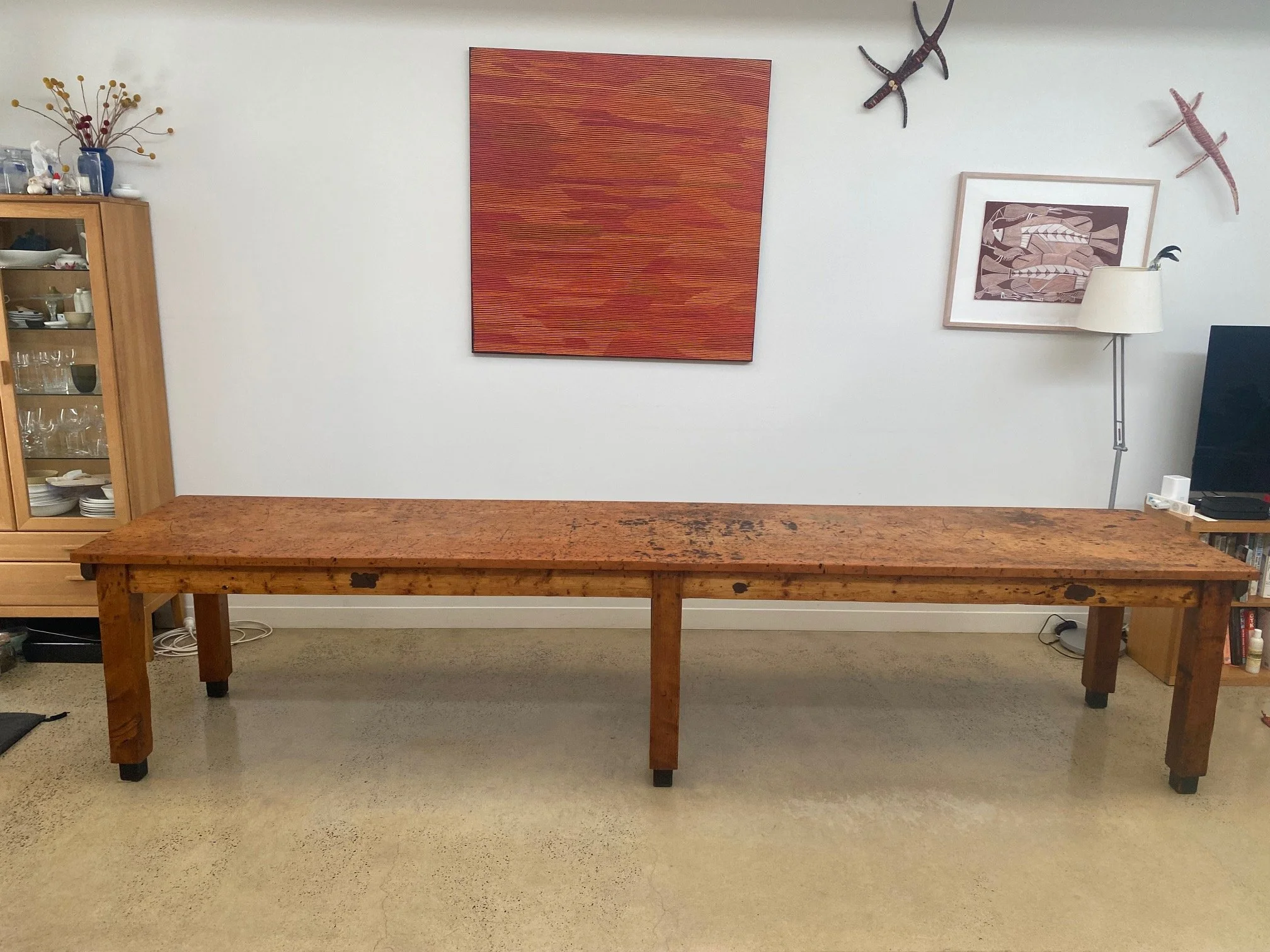 A long wooden table with a rustic appearance, placed in a room with a beige carpet and white walls. Behind the table are wall decorations including a large red abstract painting, three tribal-style wall sculptures resembling lizards or dragons, and a
