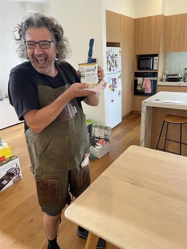 Man with curly hair and glasses smiling and holding a jar of Osmo Polyx-Oil in a kitchen.