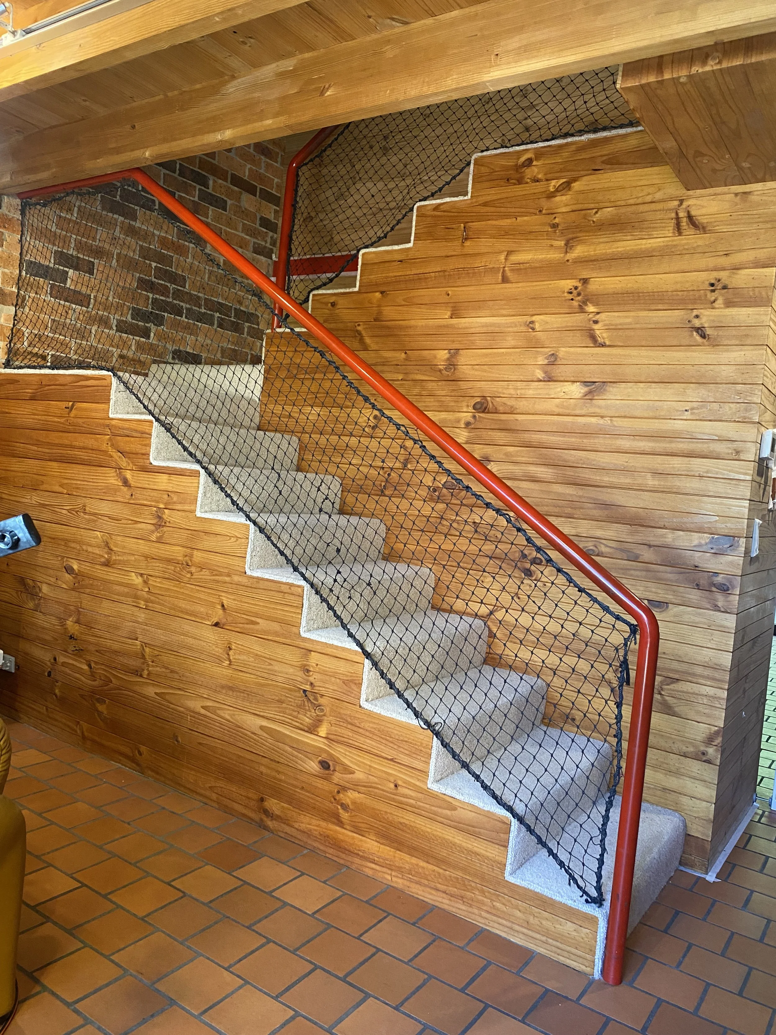 Interior staircase with carpeted steps, red handrail, wooden wall paneling, brick wall, and terracotta tile flooring.