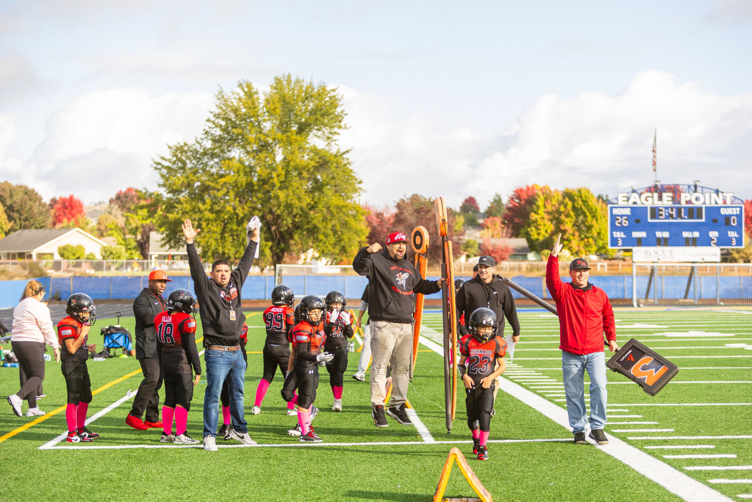 Youth football team with coaches celebrating on the field after a game at Eagle Point football stadium with a blue scoreboard in the background.