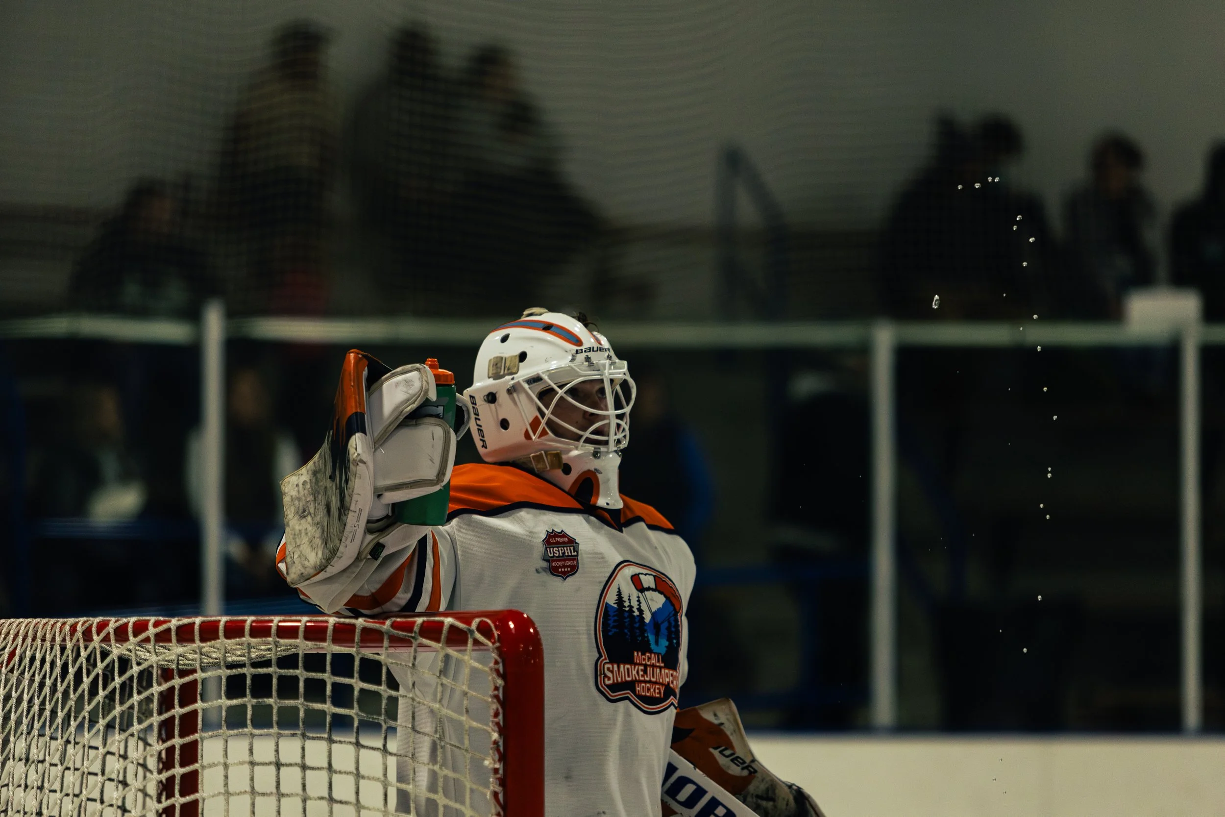 Hockey goalie in white jersey and helmet, standing in front of the net, holding a water bottle, with spectators blurred in the background.