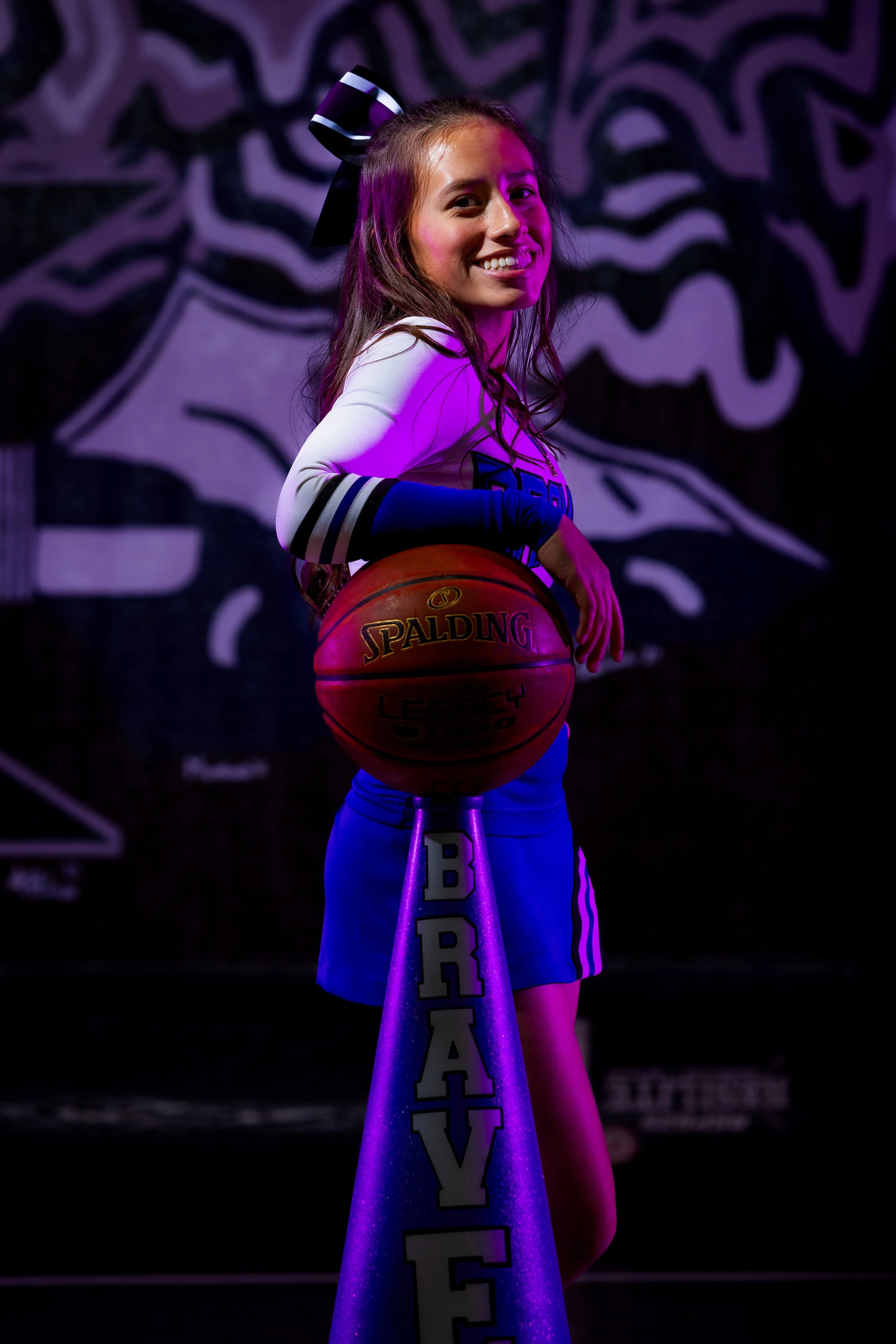 Young woman with long hair holding a basketball on a stand in a gym or sports arena, smiling at the camera, with purple and pink lighting and a stylized mural in the background.
