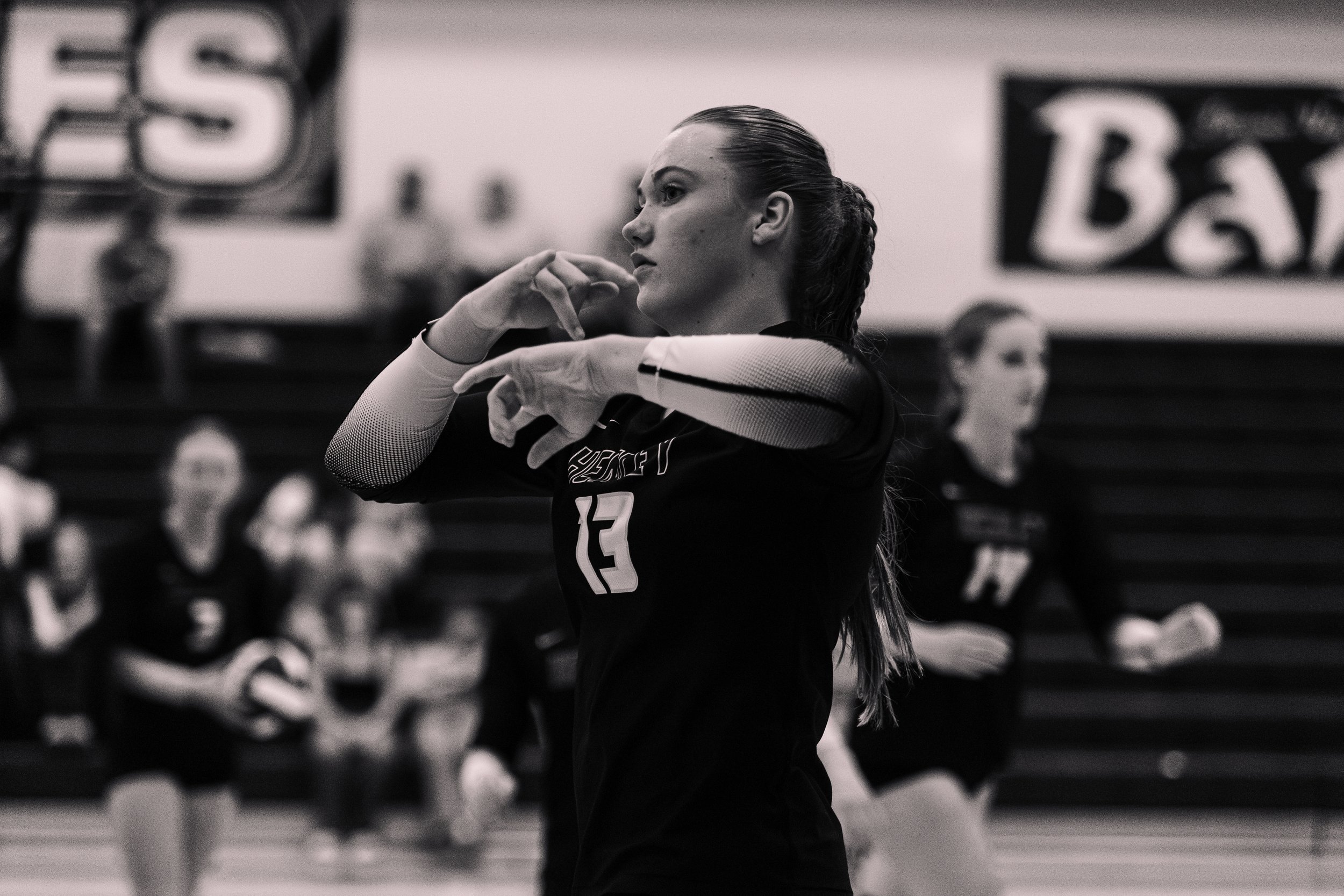 A female volleyball player in uniform adjusting her wristbands during a game, with teammates and banners in the background.