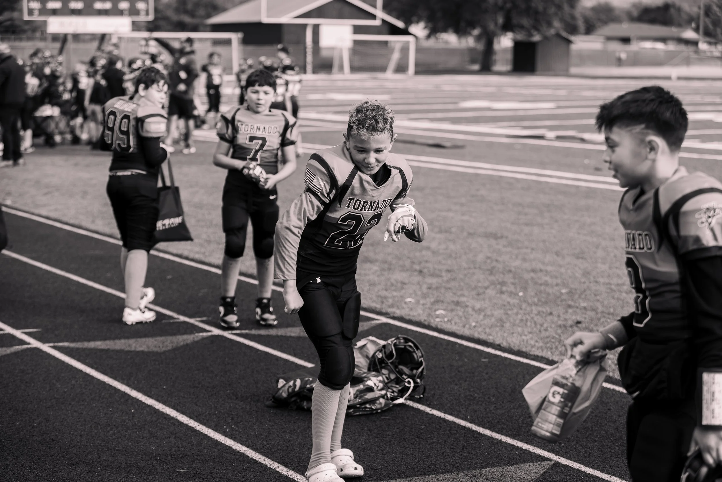 Young boys in football uniforms on a track field, preparing before a game or practice, with football gear and equipment around them.