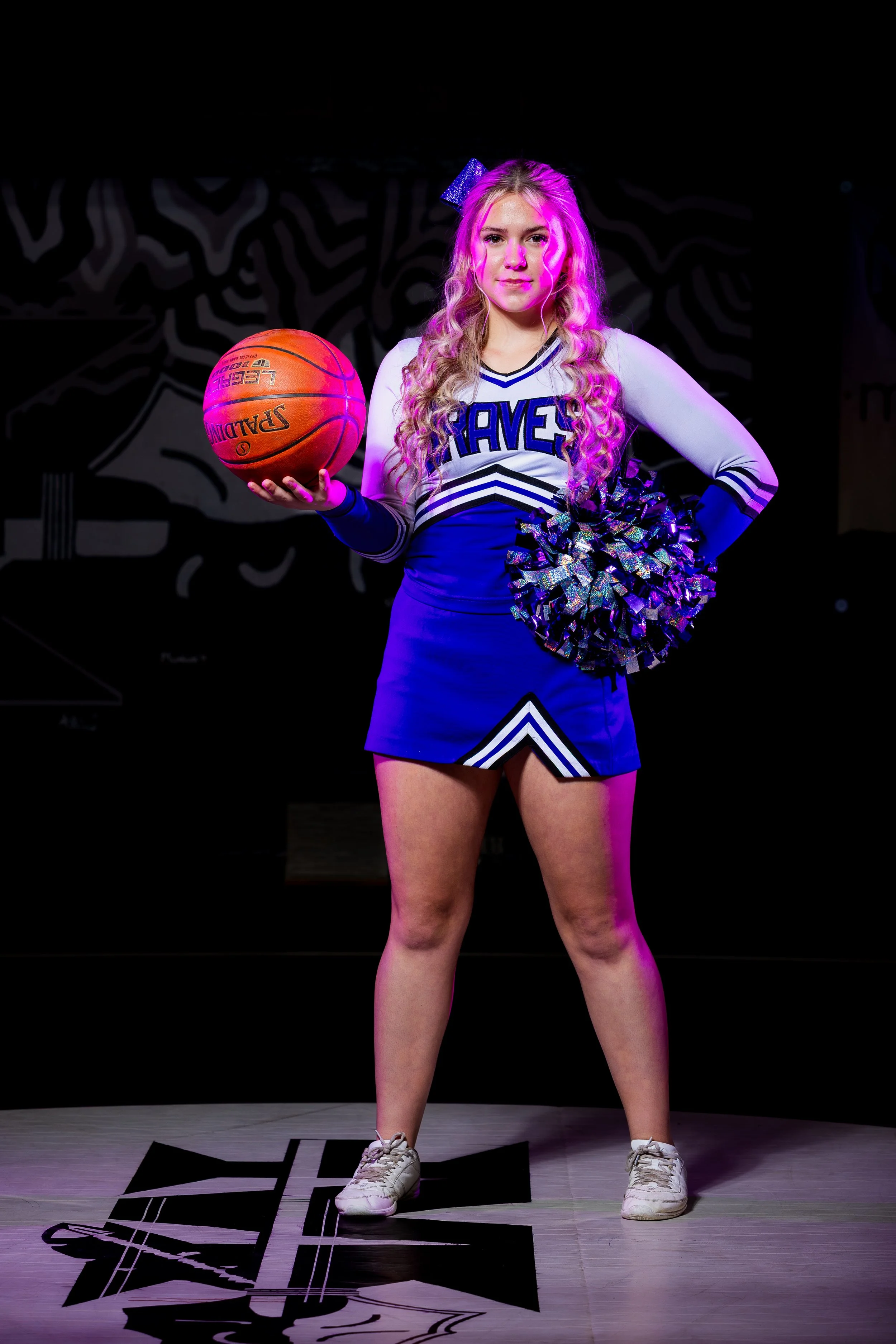 A young girl in a cheerleading uniform holding a basketball and a pom-pom, standing on a stage with a dark background and spotlight lighting.
