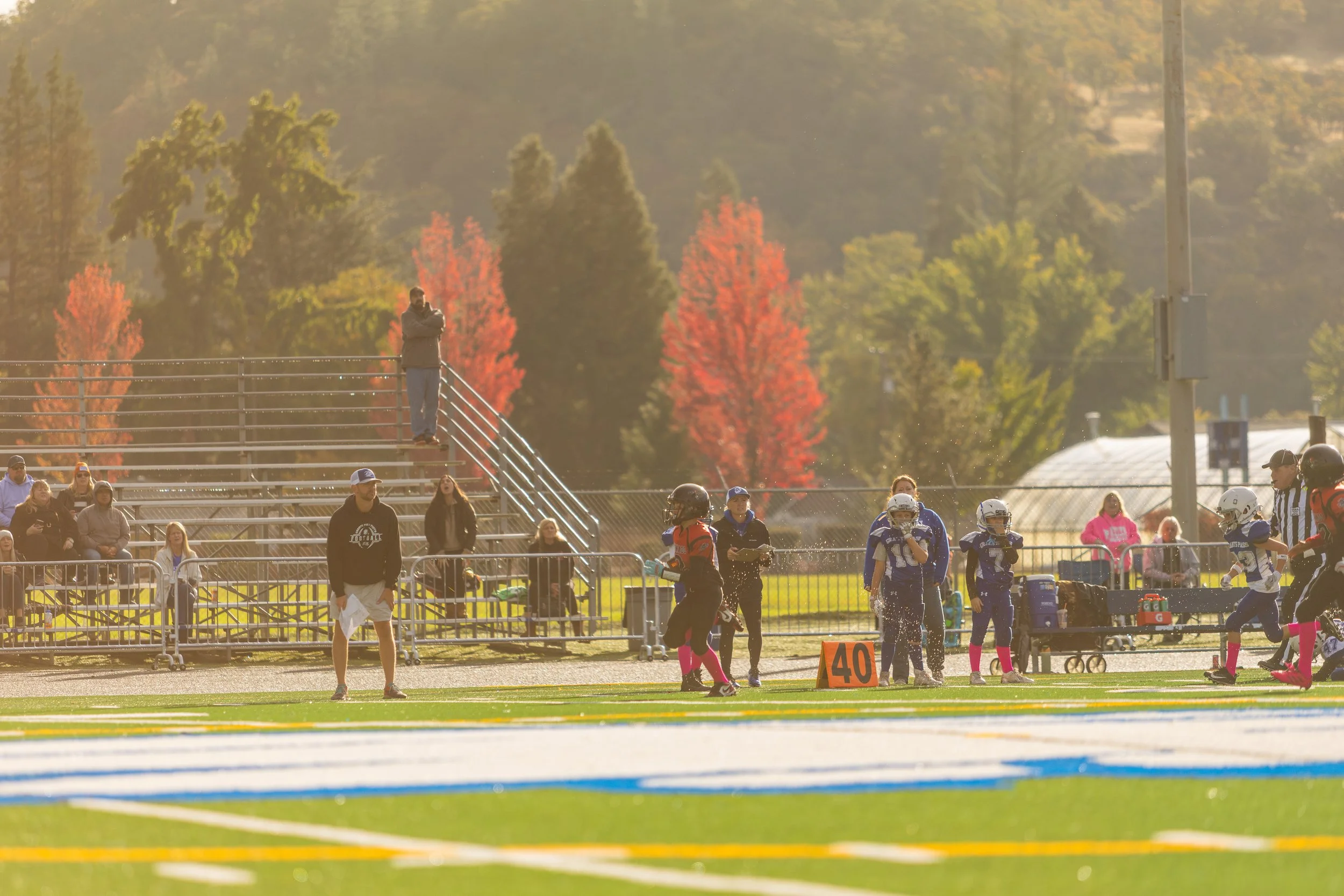 A youth football game on a bright day with players, coaches, and spectators, with colorful autumn trees in the background.