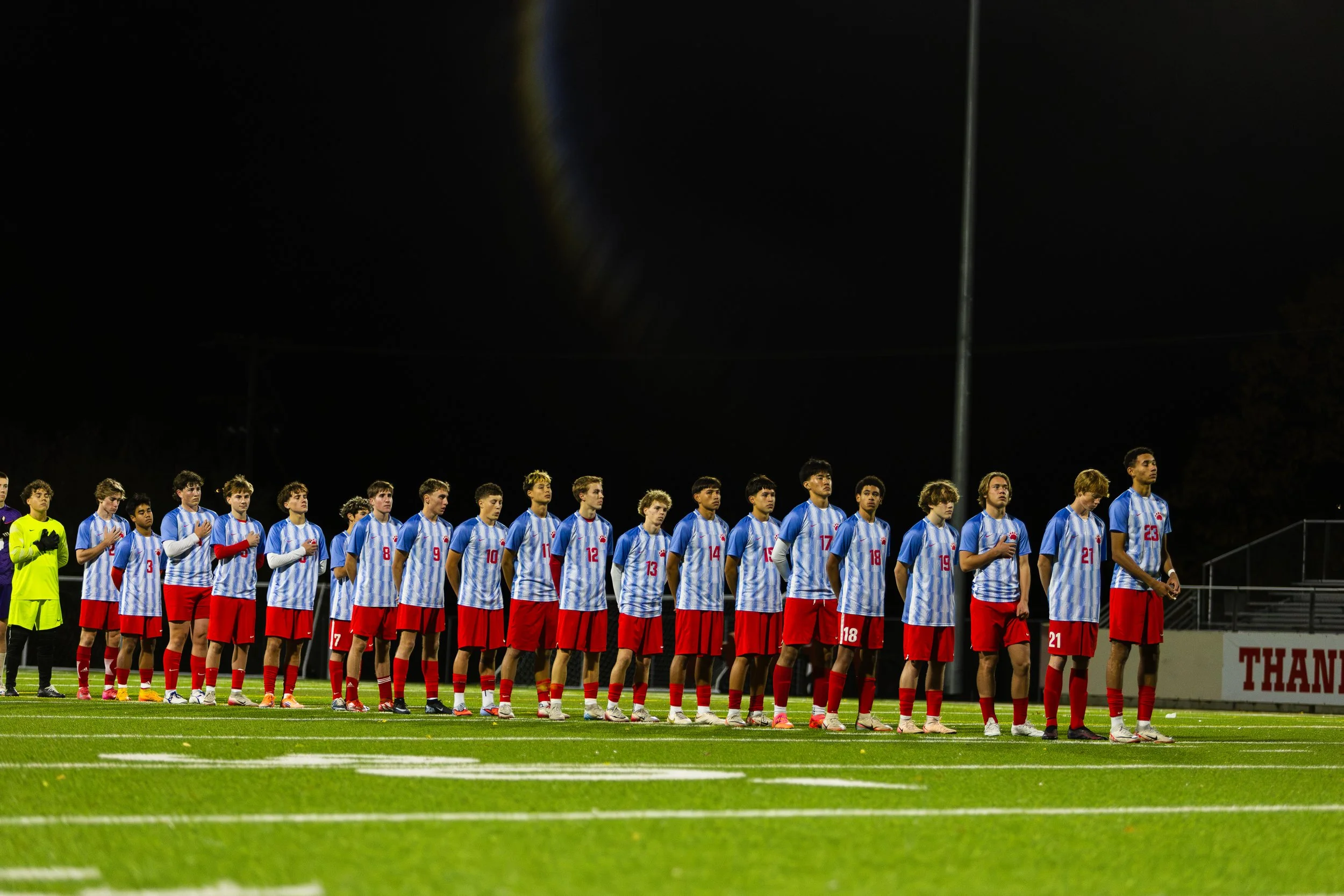 A group of young male soccer players standing in a line on a field at night, wearing matching blue and white jerseys and red shorts, with some players placing their right hands over their hearts.