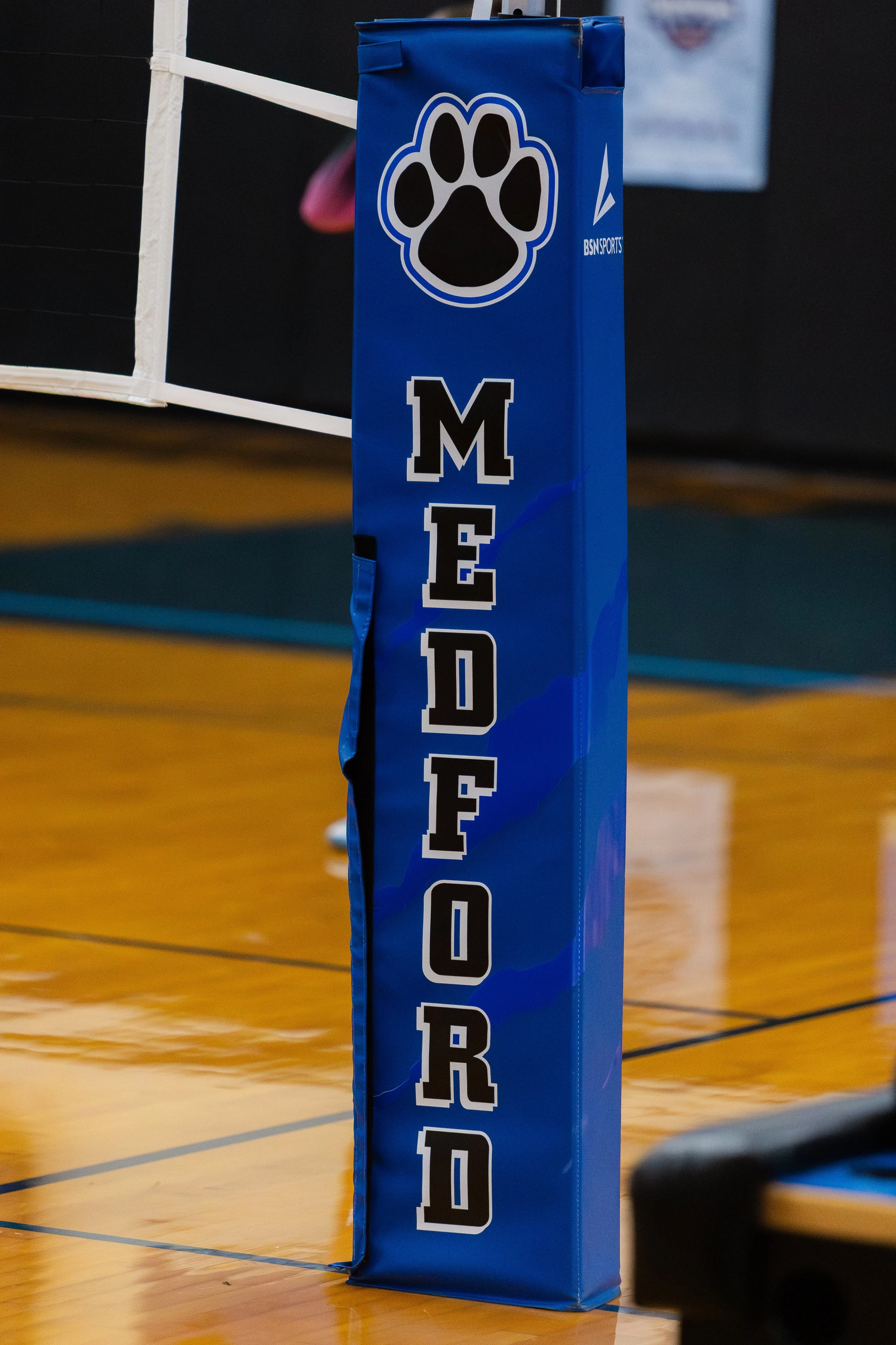 A blue padded post with a black paw print logo and the text 'MEDFORD' written vertically on it, located on a gymnasium floor.