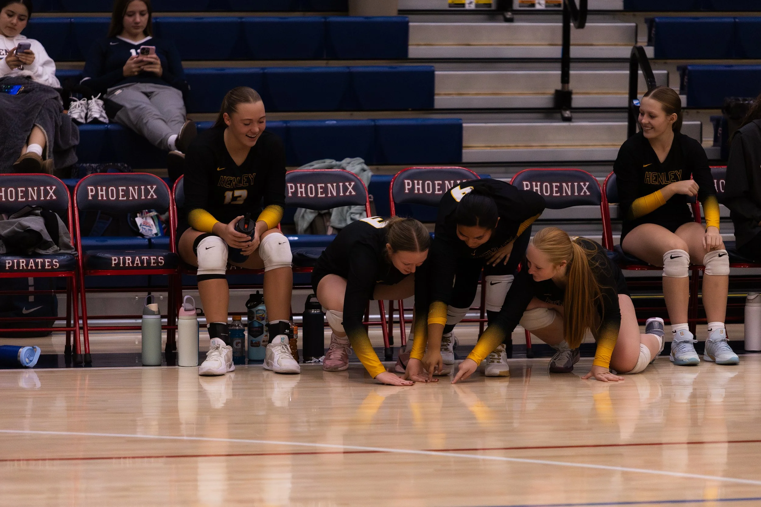 Girls volleyball players sitting on chairs on the sidelines and three girls doing push-ups on the gym floor during a game.