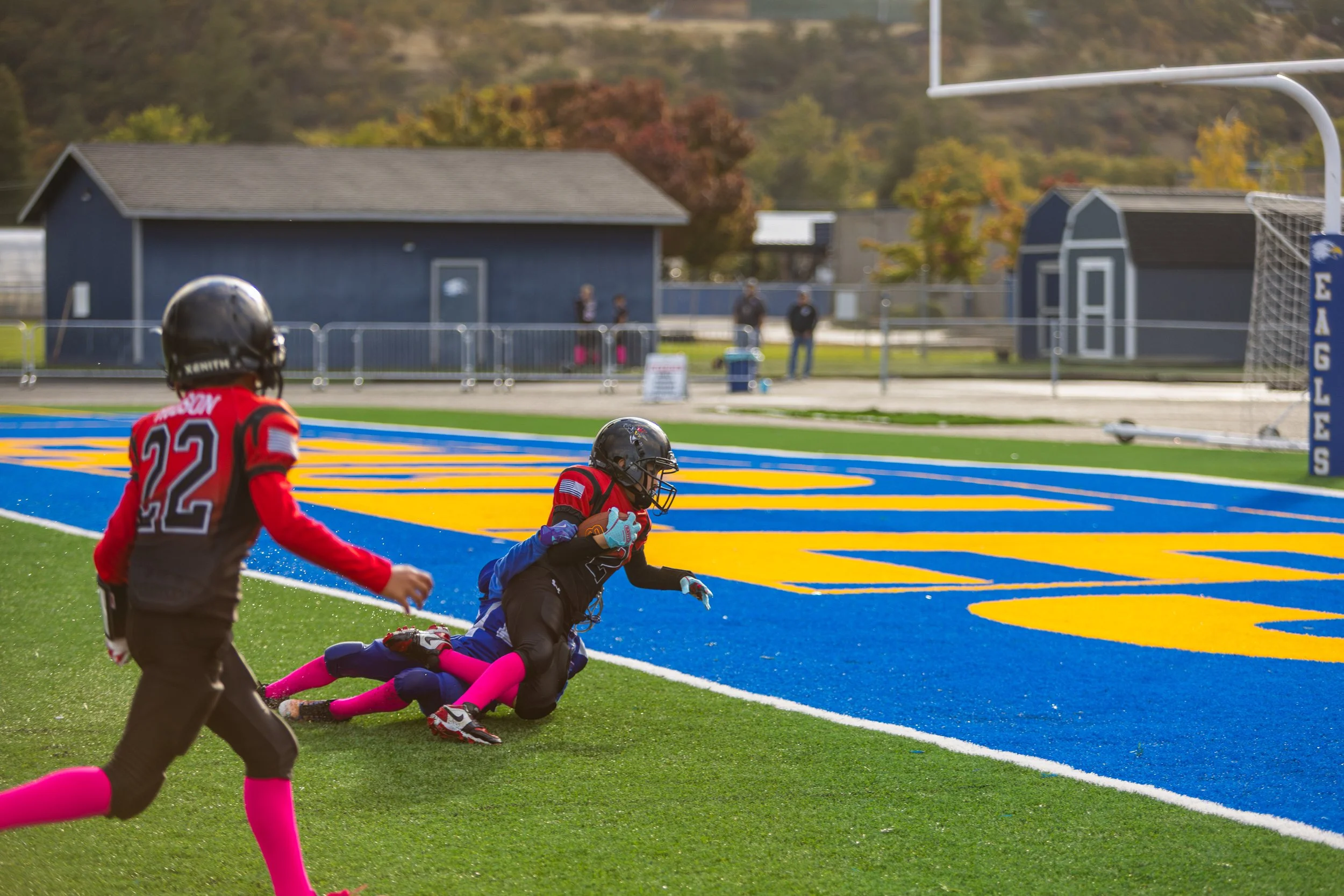 A youth football player in a red and black uniform is running with the football into the end zone, while another player in a similar uniform is on the ground in front of him. The scene takes place on a football field with a vibrant blue and yellow en