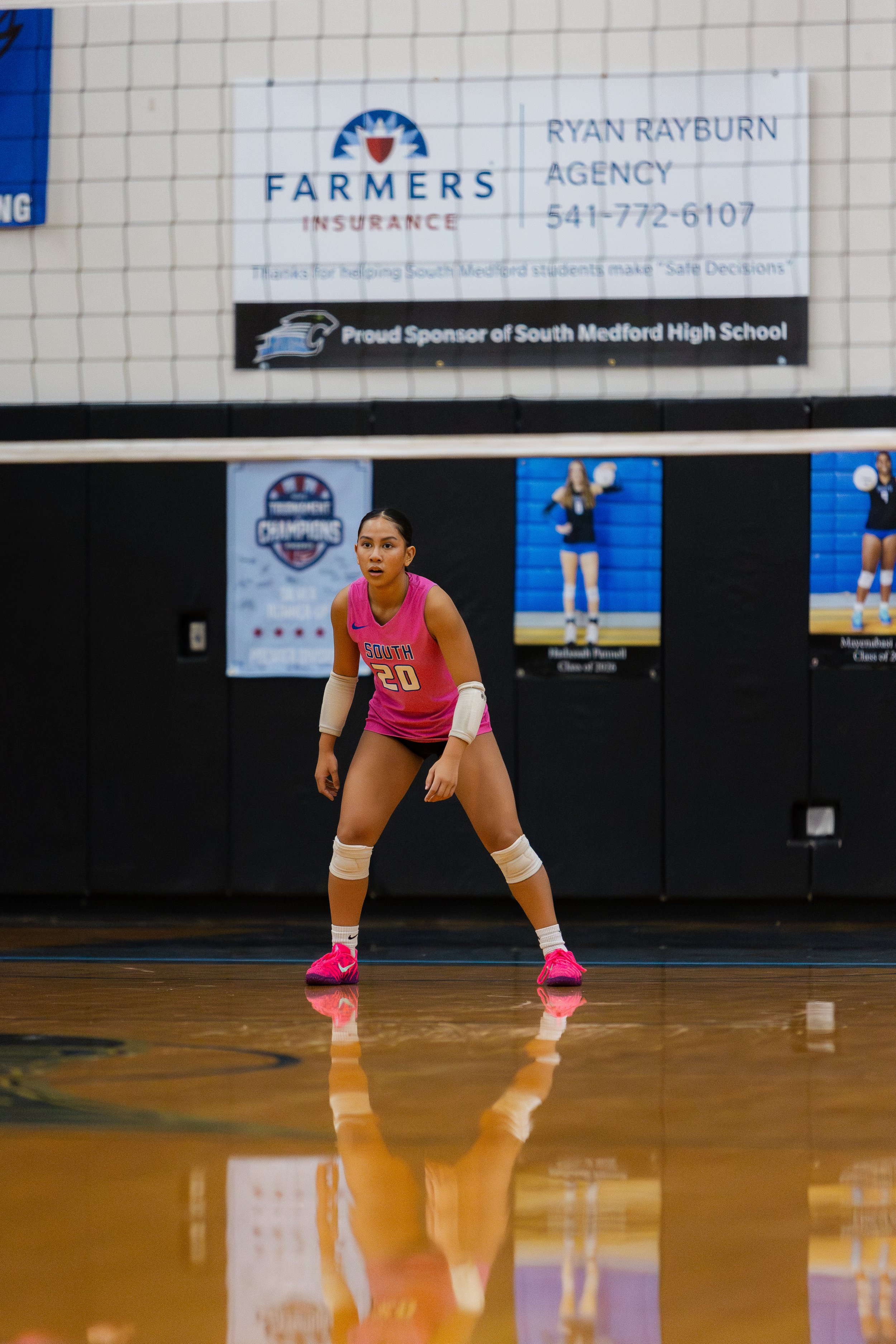 A female volleyball player in a pink uniform with the number 20, standing on a gymnasium floor, preparing to receive or set the ball, with her hands in front of her and knees slightly bent.