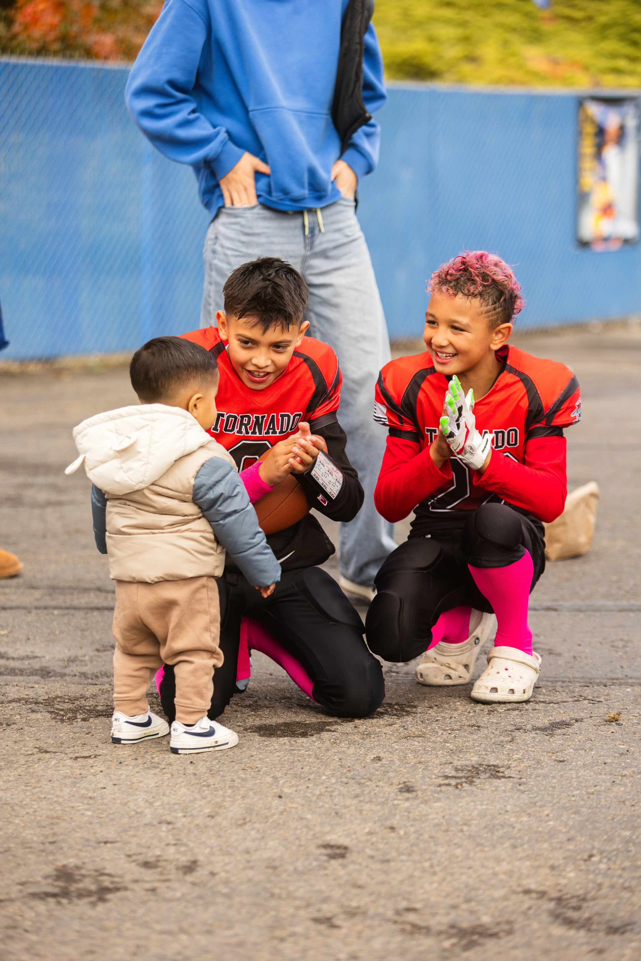 Two young football players talking to a small child, all wearing football uniforms with 'Tornado' on the jerseys, on an outdoor field with a blue fence in the background.