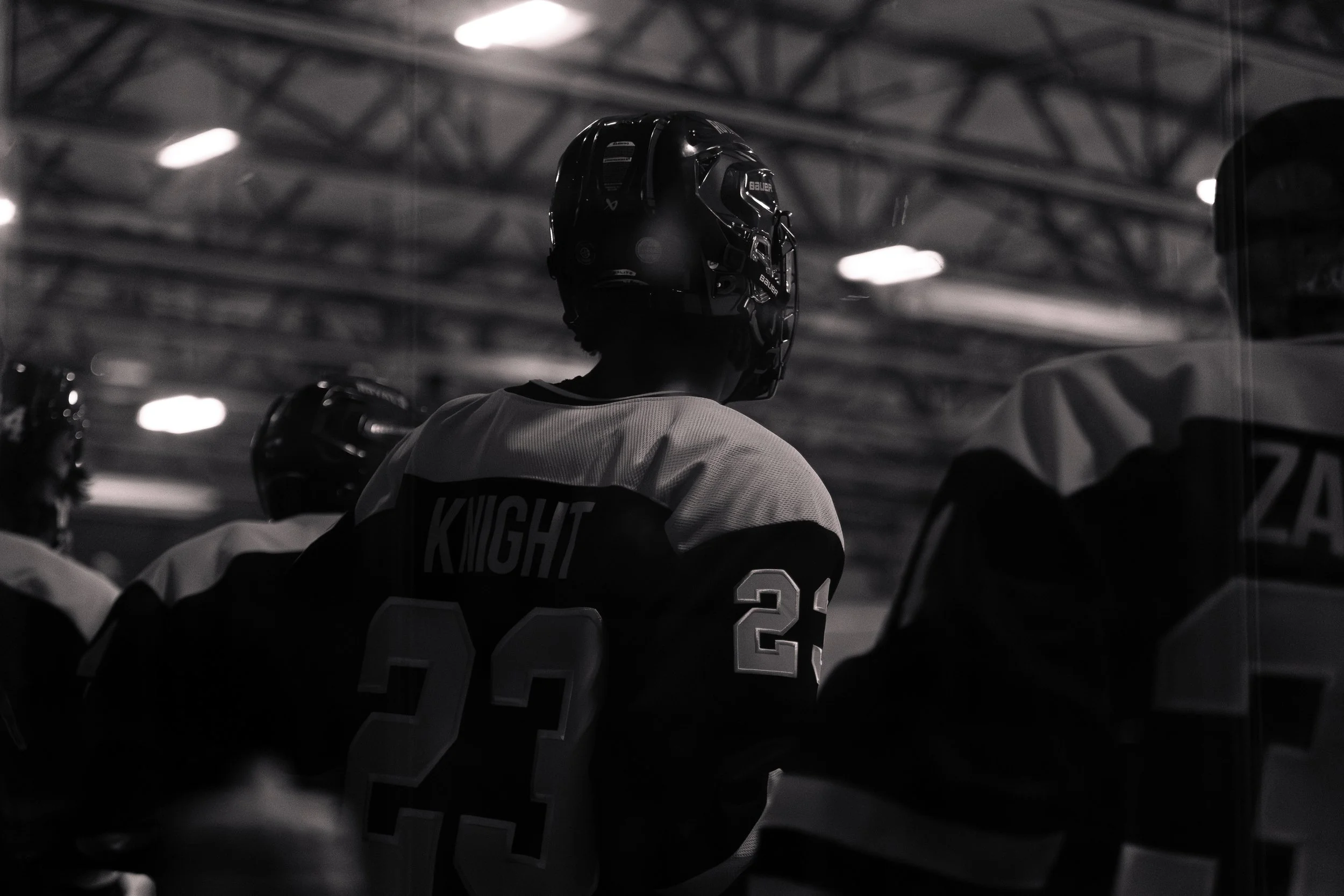 A black and white photo of ice hockey players on the bench in an indoor rink, one player wearing a helmet and jersey with the name "Knight" and the number 23.