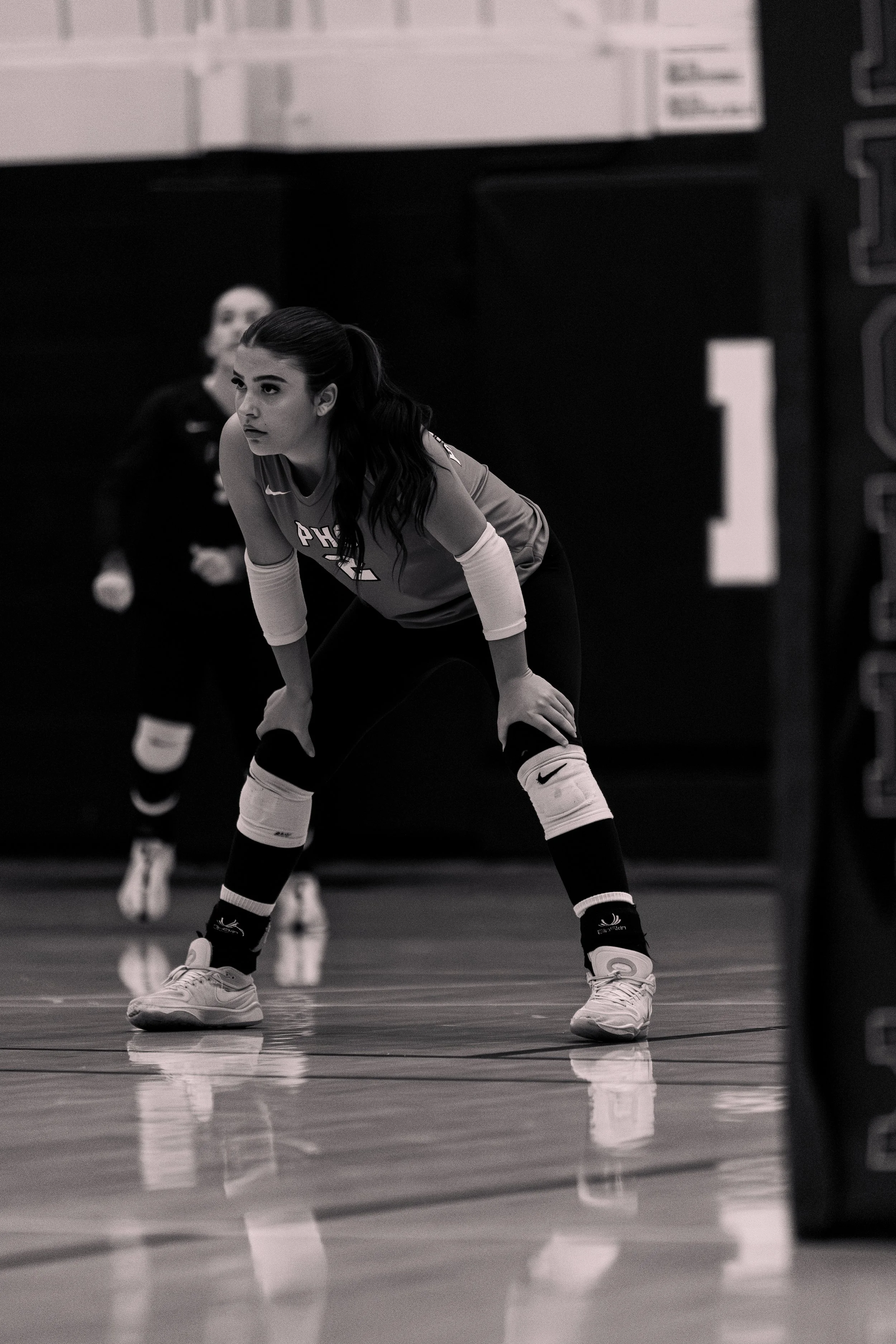 A female volleyball player in a ready stance on the court, with another player in the background, in a gymnasium.